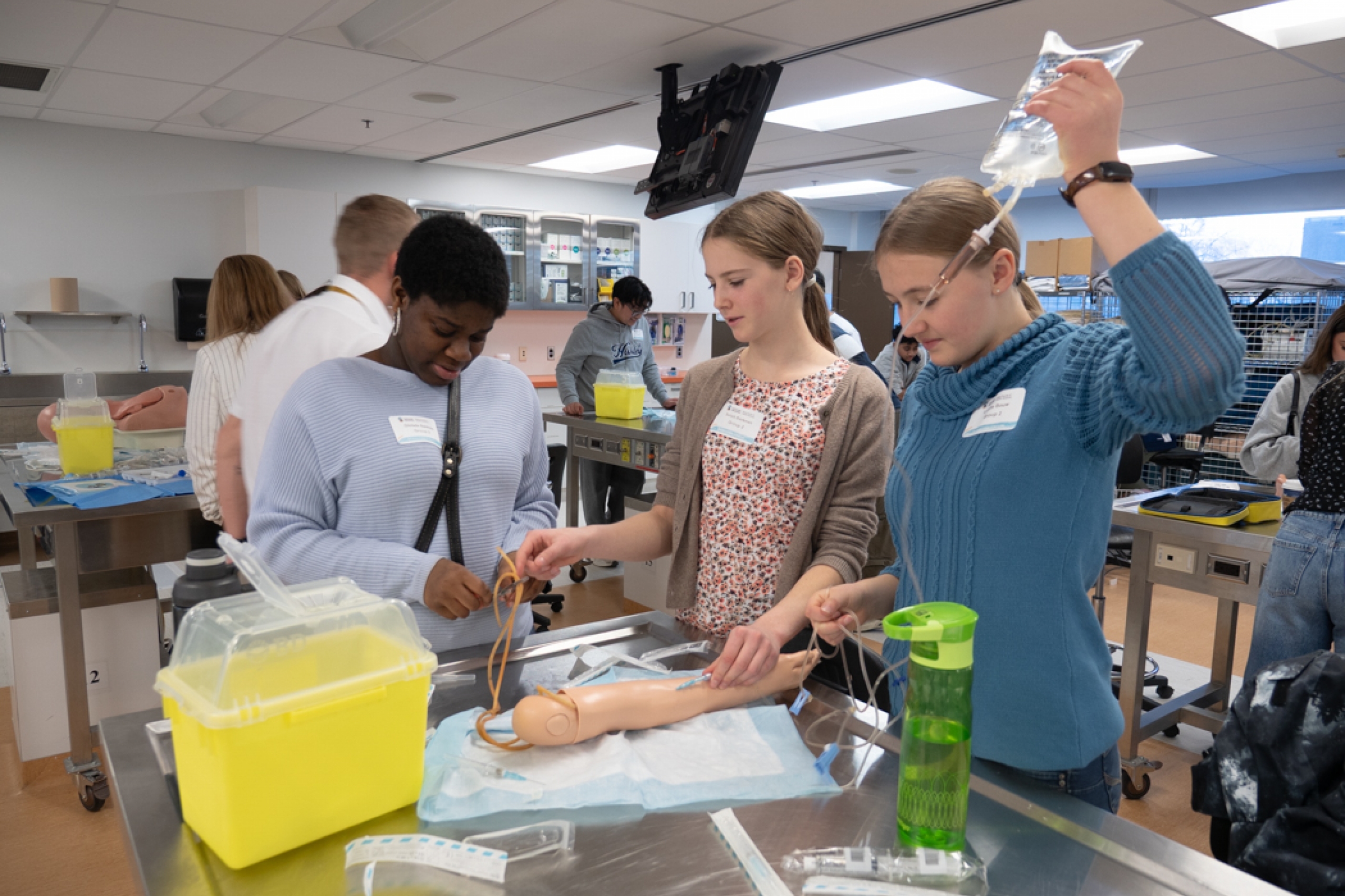 Three students in a clinical skills session: two practice on a manikin arm while another holds an IV drip connected to the manikin.
