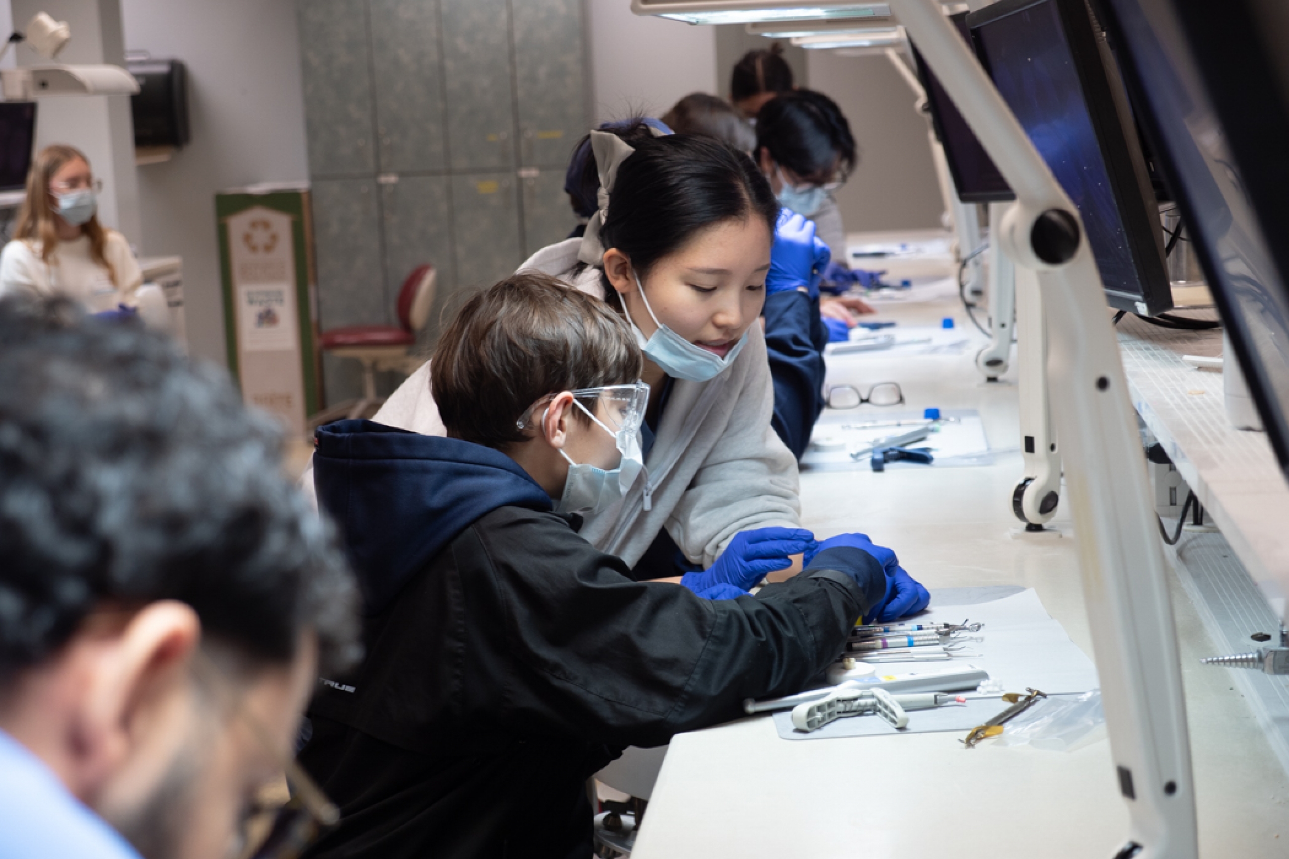 In a dental school lab, a dental student assists a Grade 9 student in practicing a tooth filling on a model.