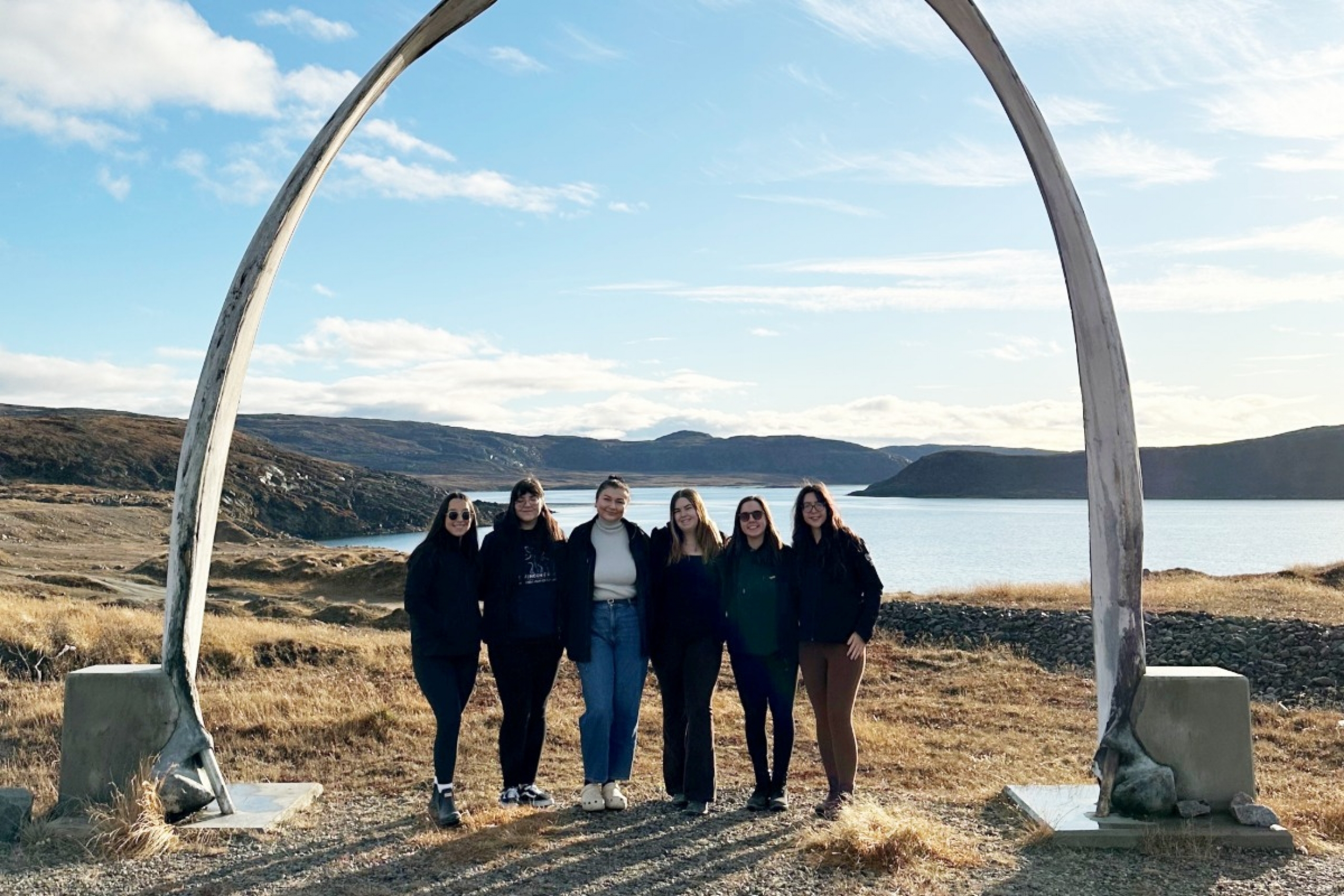 group of students stand and smile beneath whale bone arch with northern tundra in background.