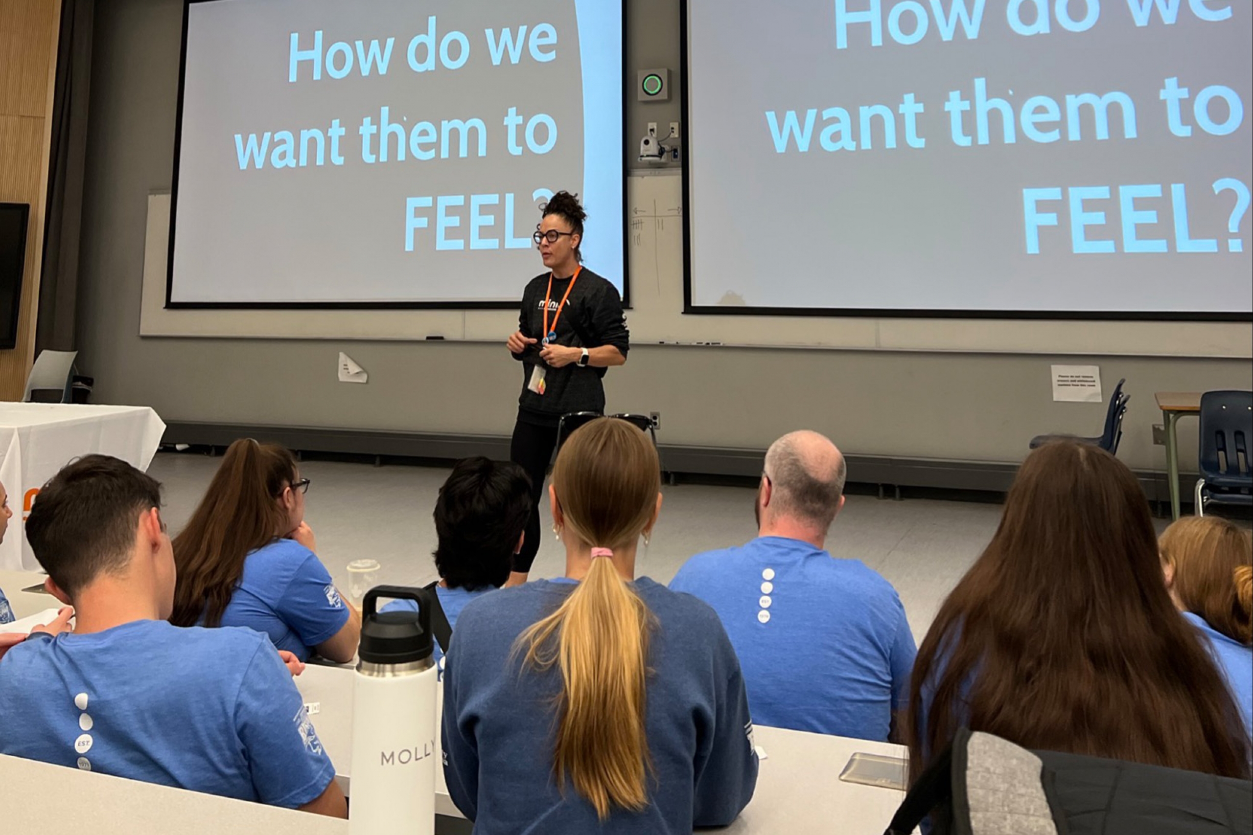 Ashley Gagnon stands in front of a lecture hall facing a group of young people in matching blue shirts. The screen behind her reads: How do we want them to feel?