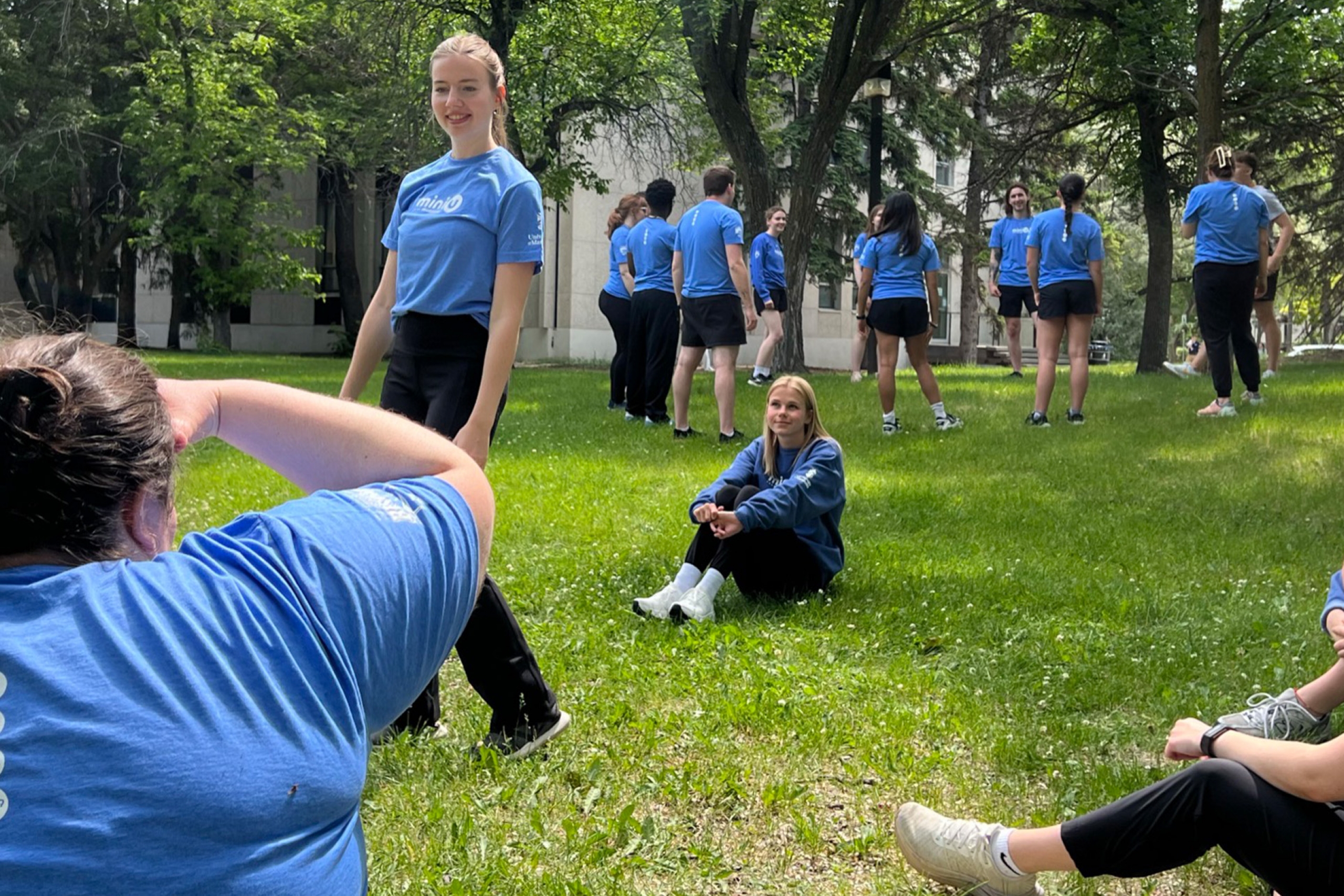 A group of young adults in matching blue shirts stand and sit on a green lawn during Mini U leadersweek in 2025.
