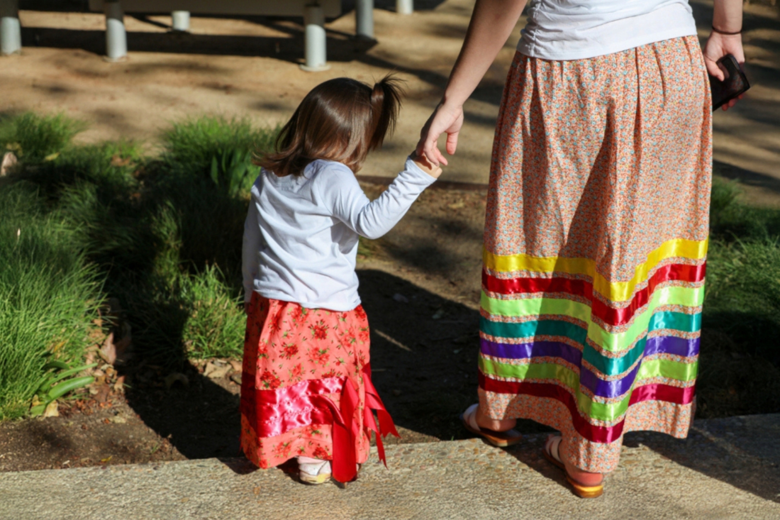 An adult is holding a child's hand. They are wearing ribbon skirts.