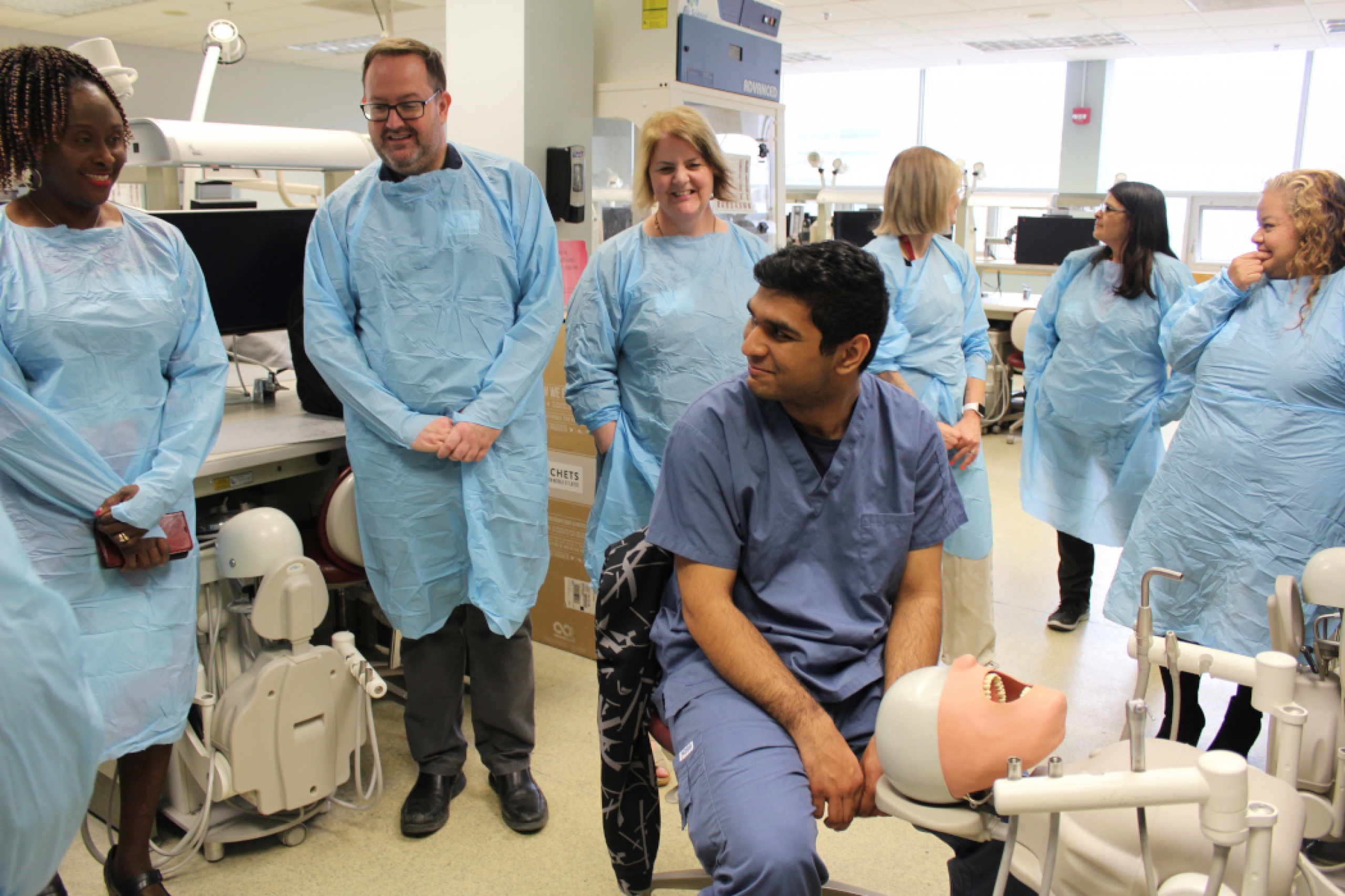 Six people wearing medical gowns stand around a student seated in front of a dental manikin.