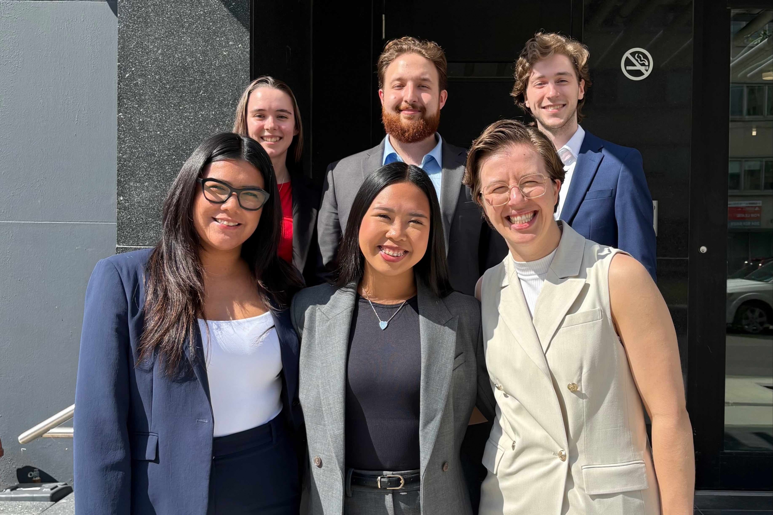six law students in suits smile in the summer sun on the steps of the legal aid Manitoba clinic