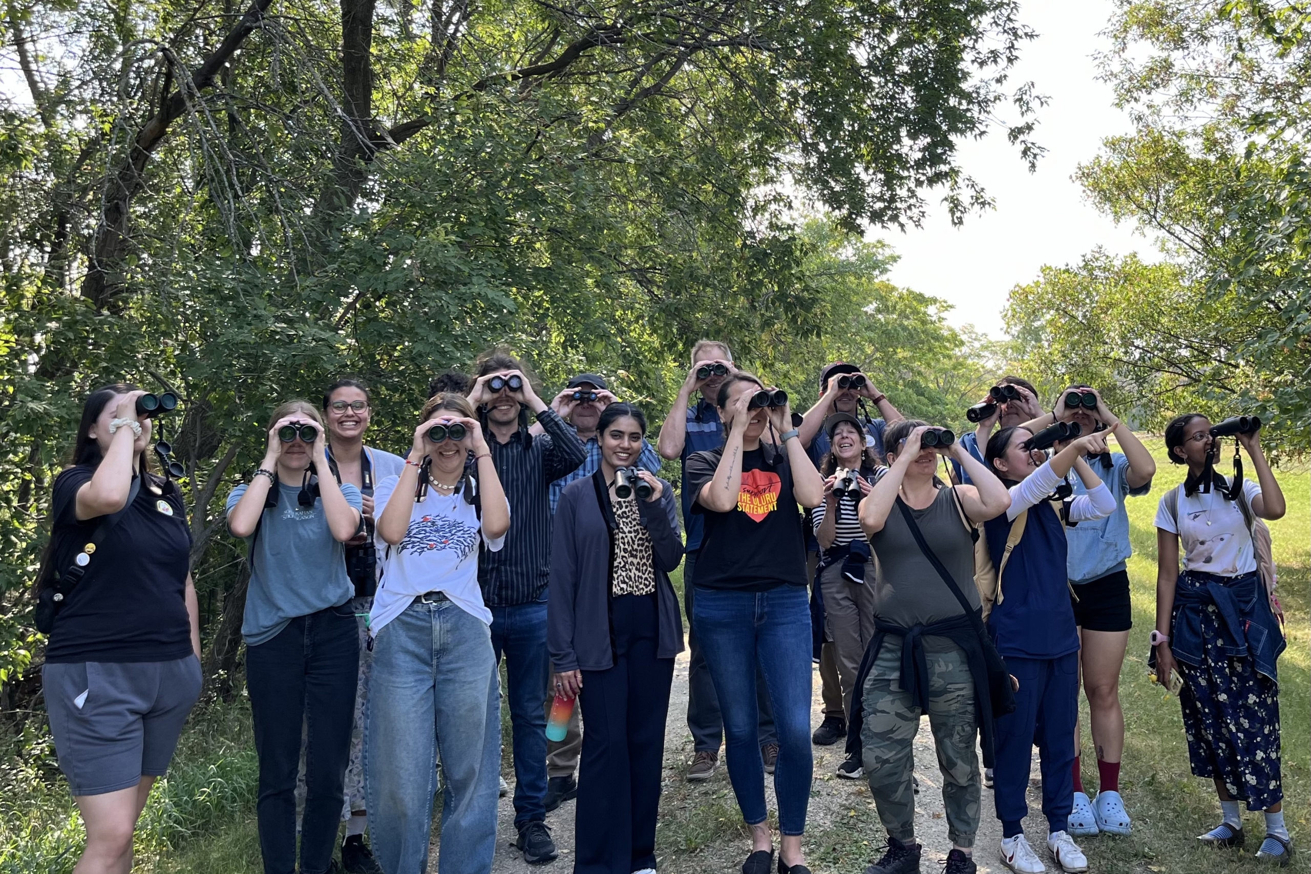 group of people holding up binoculars to their eyes and smiling, with trees in the background.