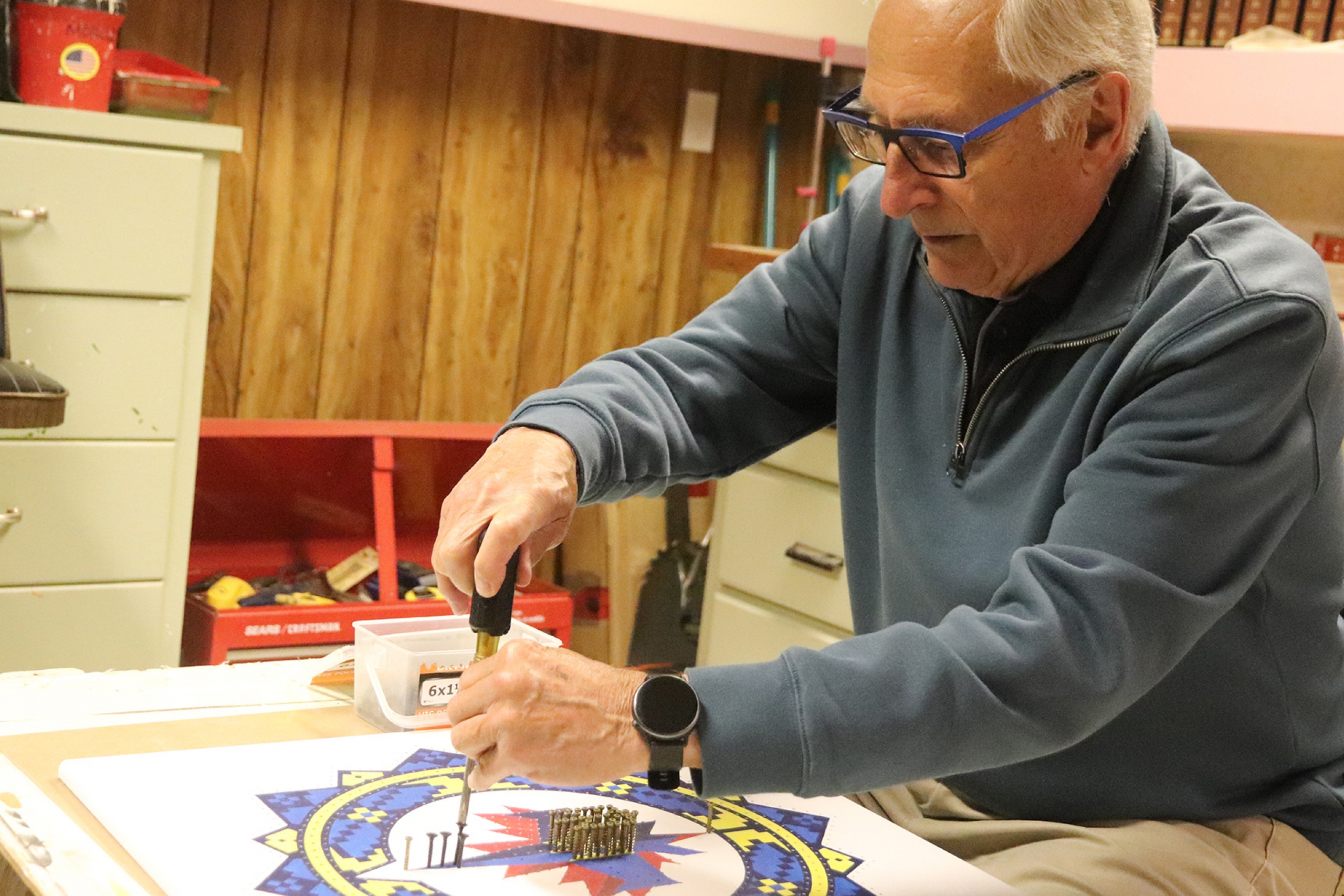 A man sits at a workbench manually adding screws to a pattern of the Winnipeg Jets Ukrianian heritage logo.