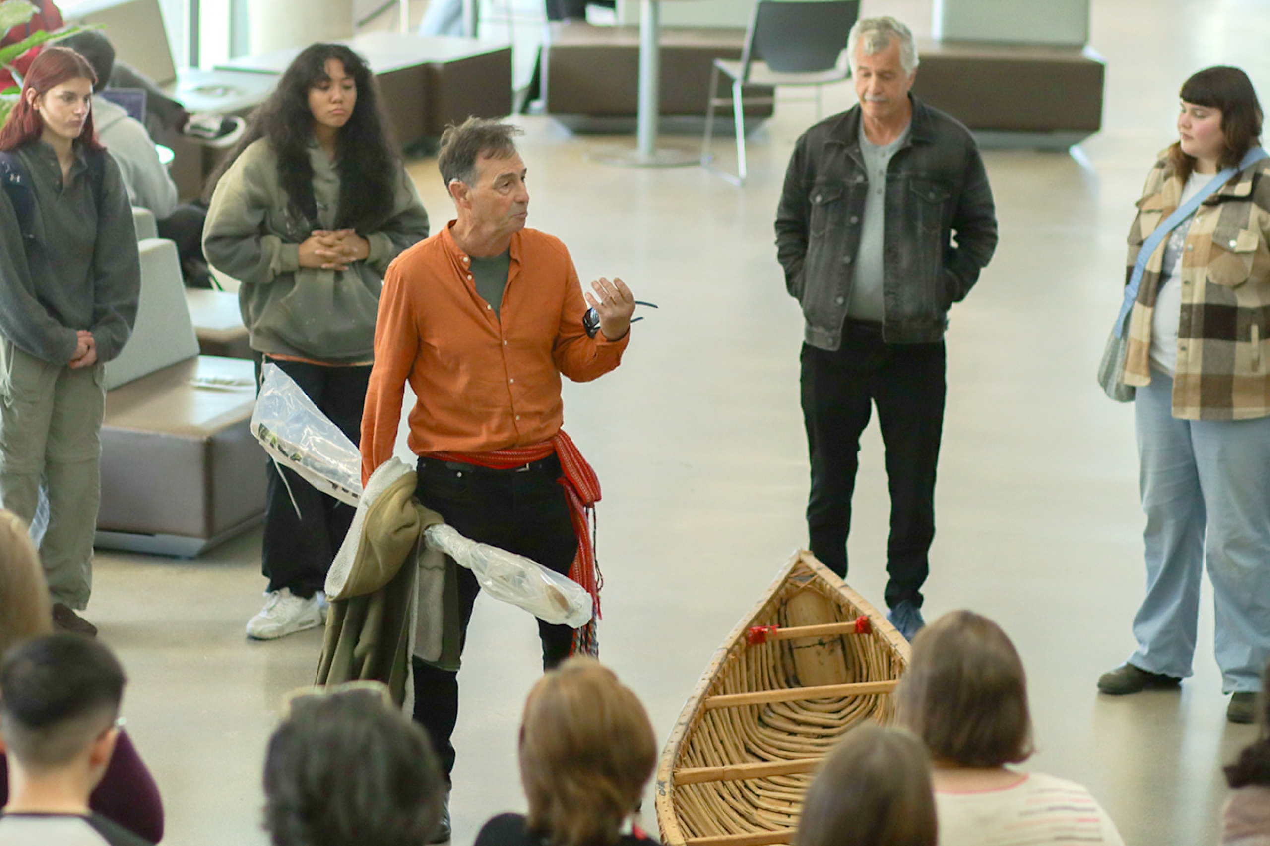 A man in an orange shirt wearing a Metis sash stands next to a handmade canoe surrounded by onlookers.