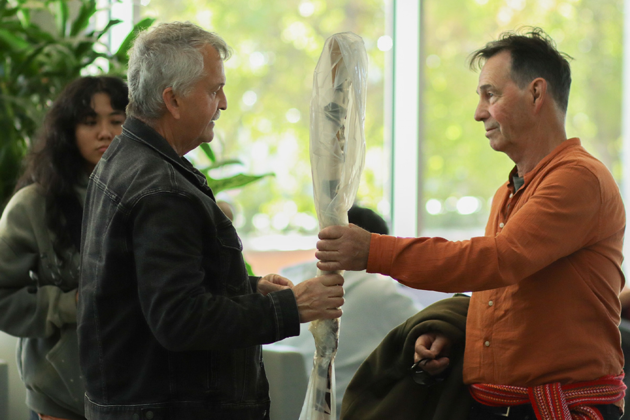 A man in an orange shirt hands a wrapped paddle to another man as part of ceremony honouring a traditional birch bark canoe project.