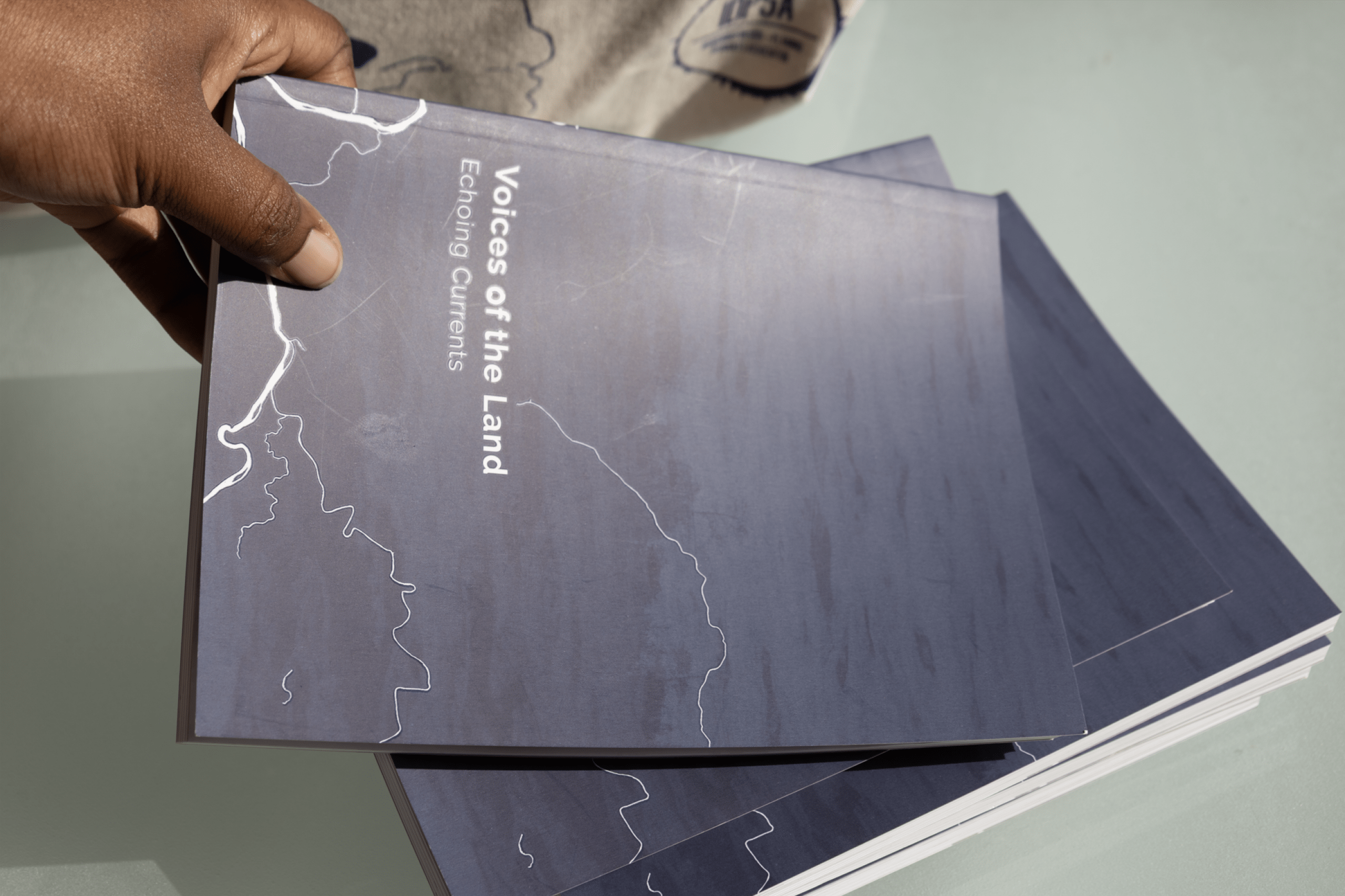 A hand holds “Voices of the Land: Evolving Currents,” a dark blue book with white map-like lines, resting atop a stack of copies. Photo by Ayla Simone