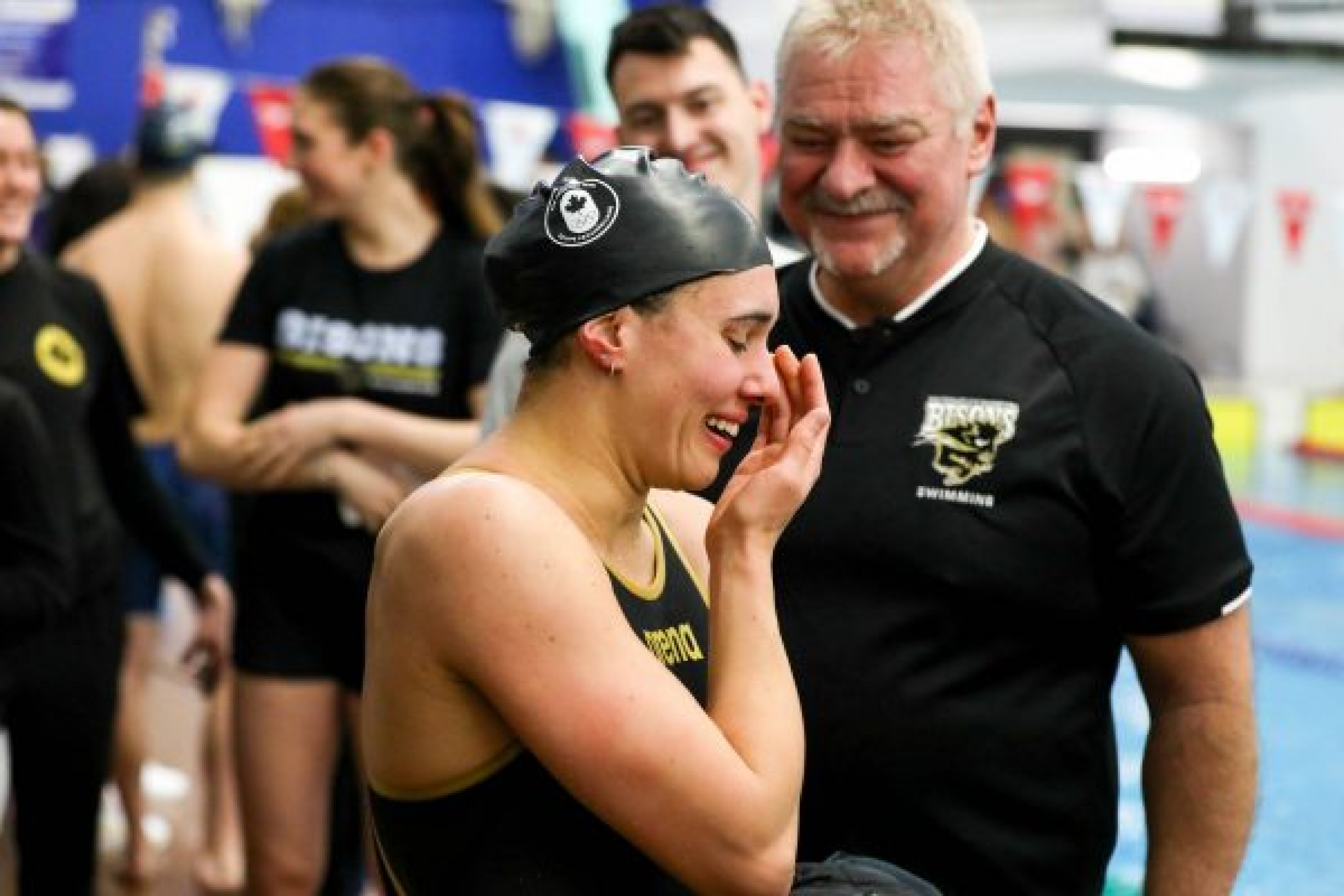 A swim athletes wipes her eye after a race with her coach looking on.