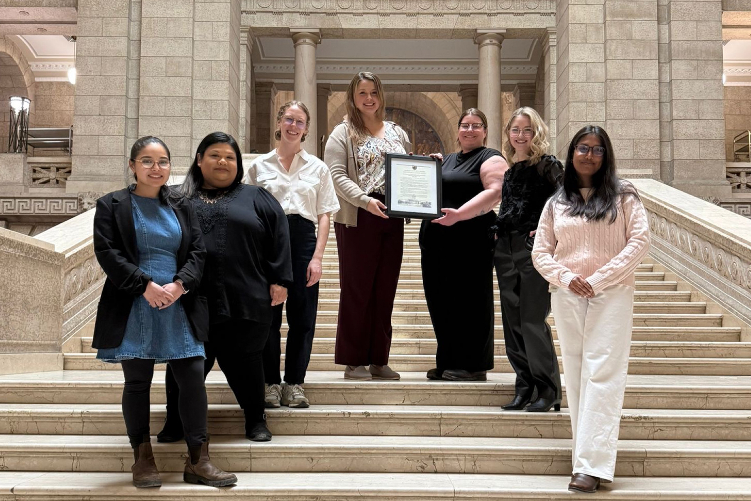 A group of students from the WISDOM program stand inside Manitoba legislative building holding a framed copy of a member's statement.