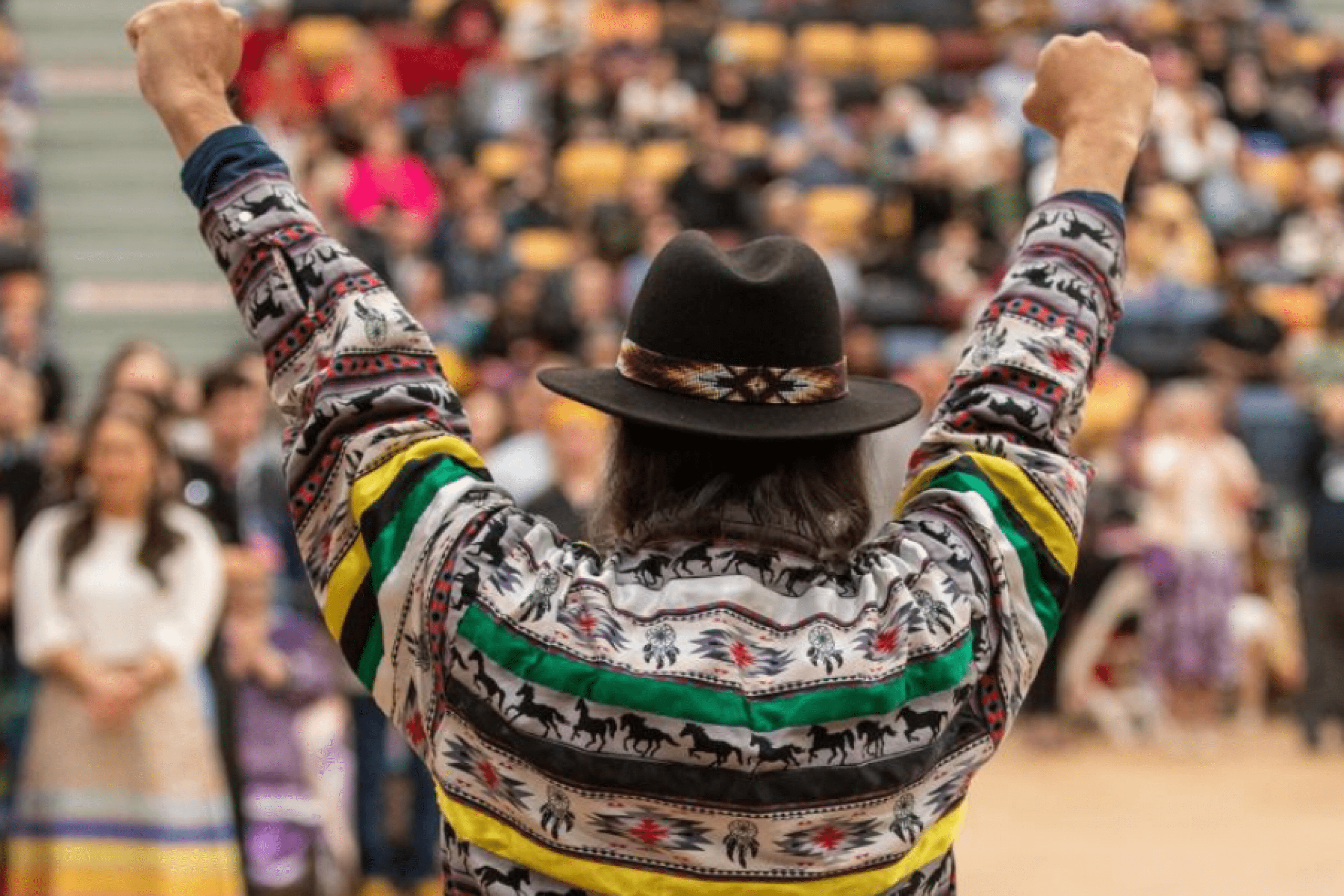 An Elder raises both arms while facing a large audience during an Indigenous community gathering.