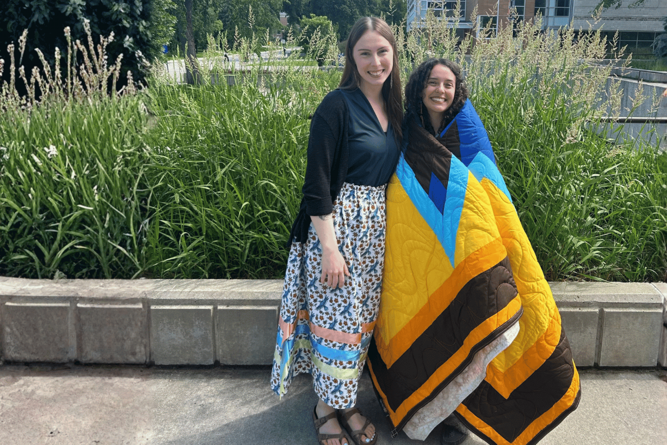 1. Bailey Pigeau stands with another person wrapped in a colourful quilt outdoors.