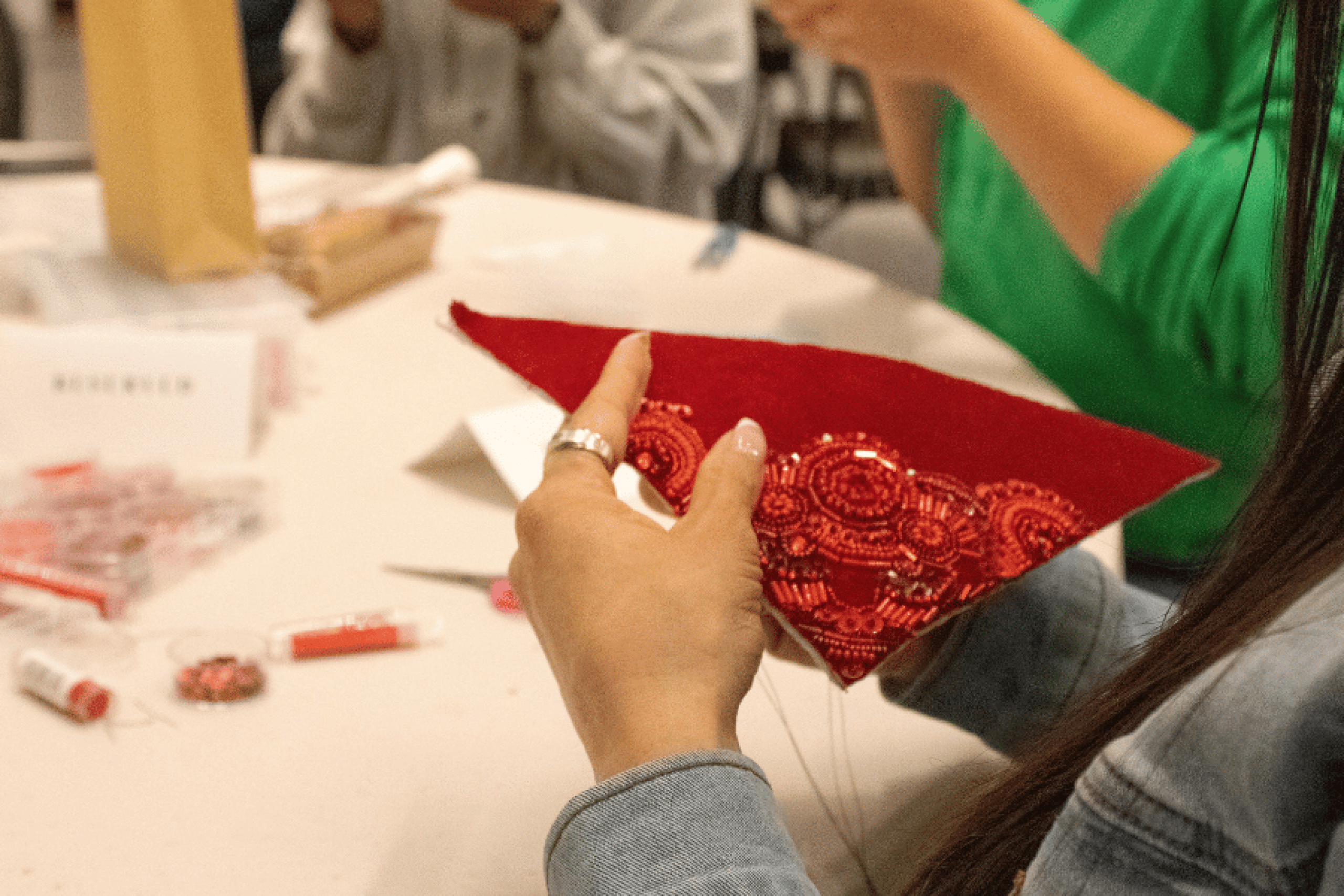 A woman holds a partially completed red beadwork piece while sitting with other participants at a table.
