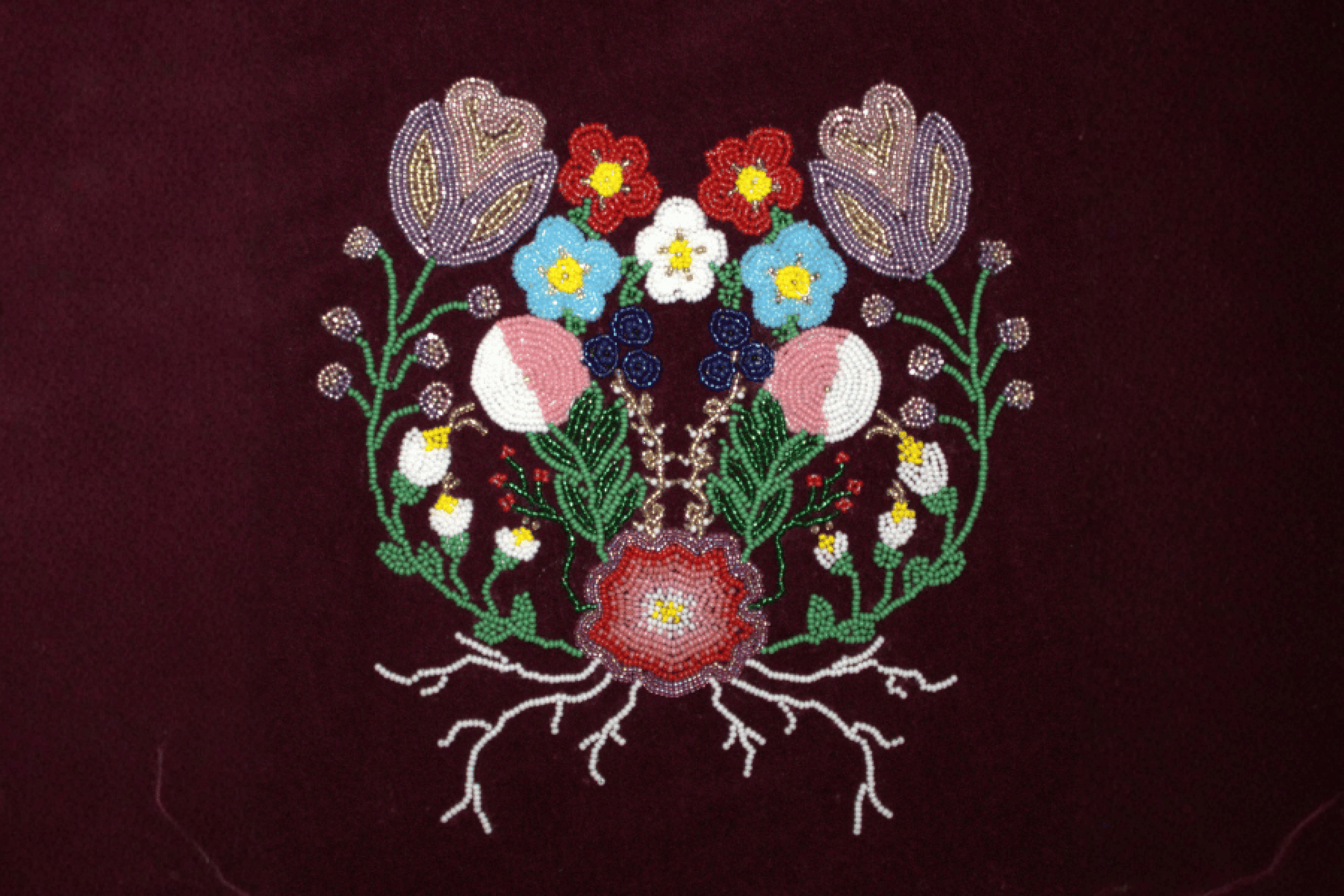A symmetrical floral beadwork design with colourful flowers, green stems and white roots on dark red fabric.