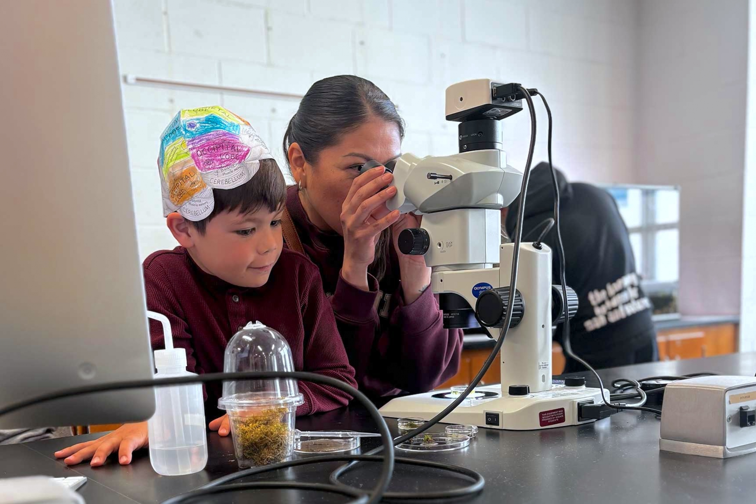 A person looking through the microscope beside a kid wearing a paper hat.