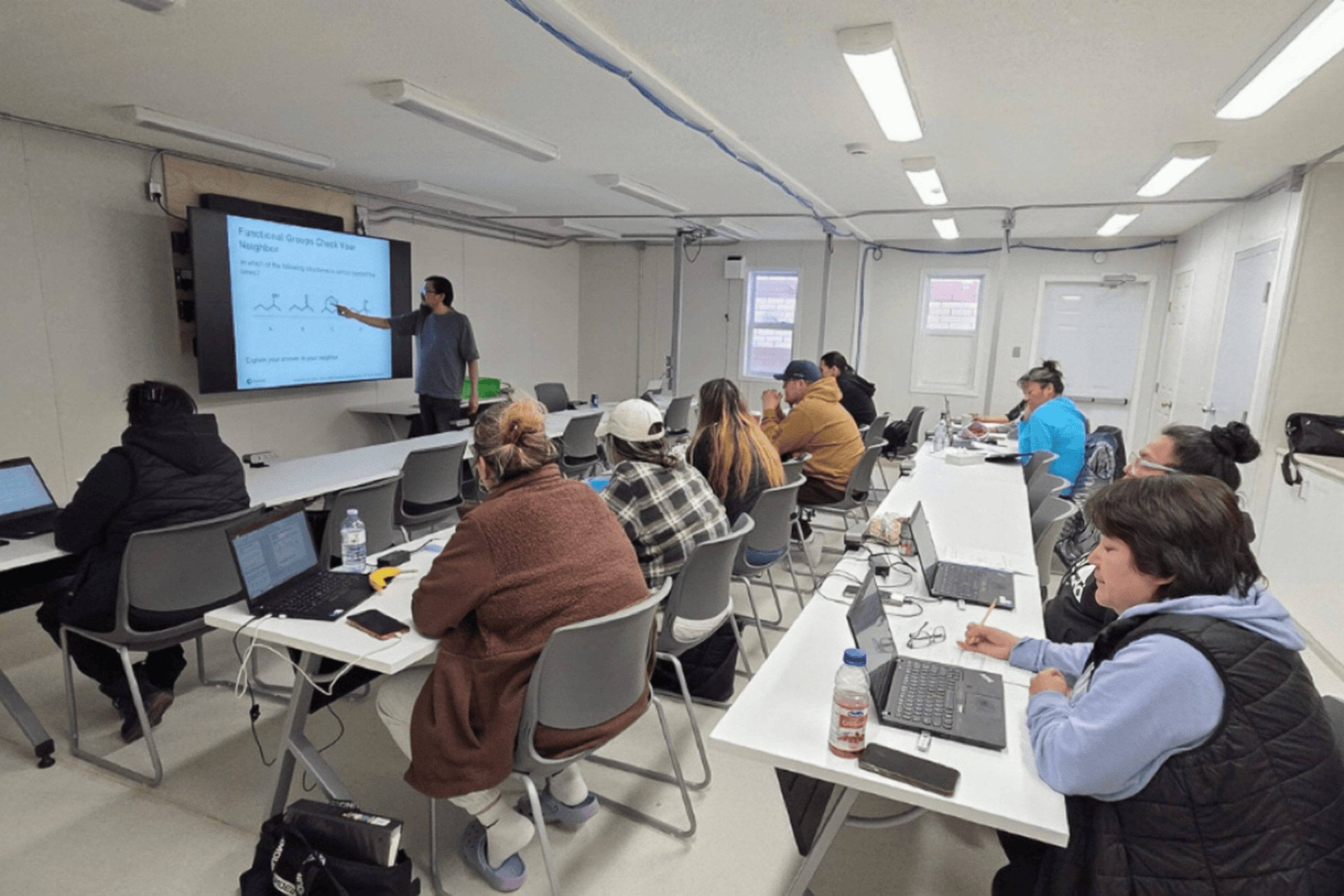 Instructor teaching in a multimedia classroom while adult students follow along on laptops.