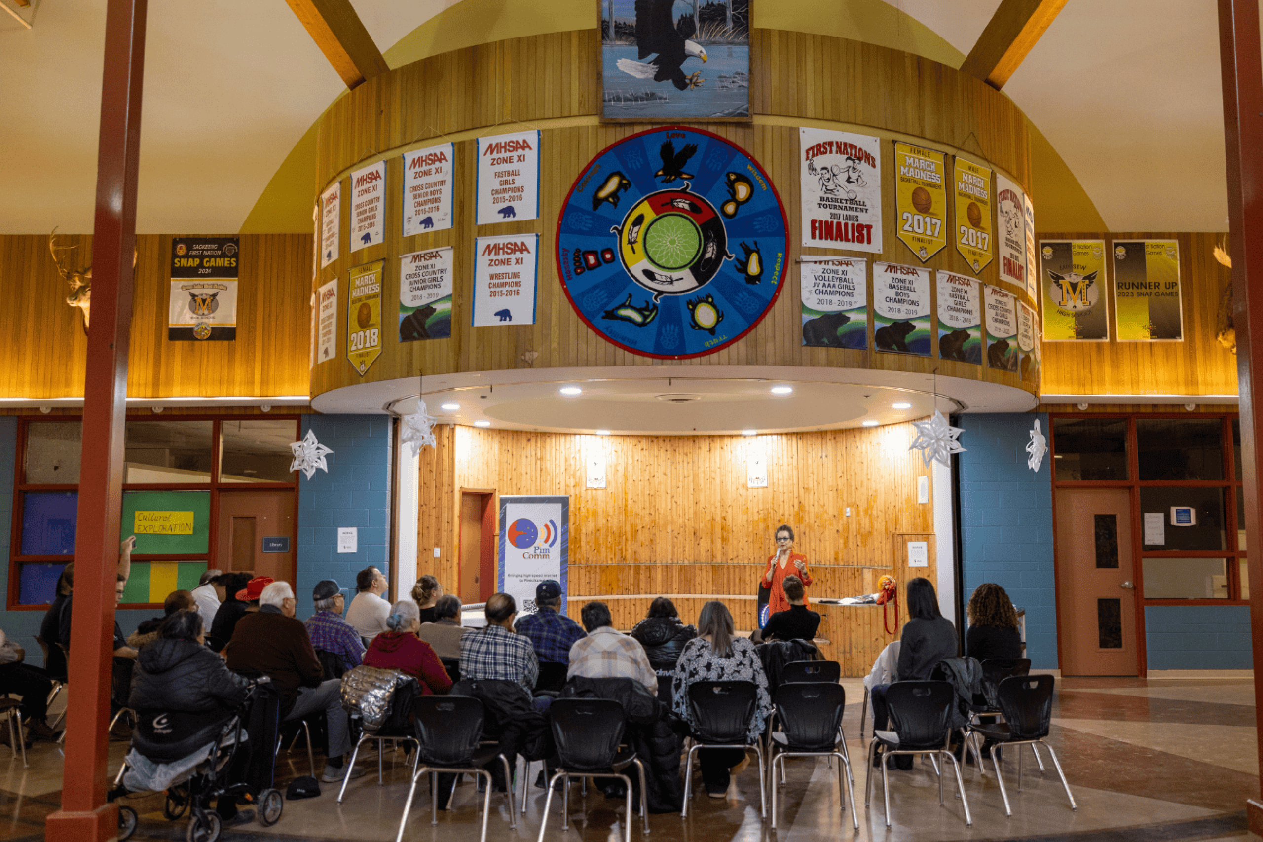 Audience seated in a school hall listening to a speaker beneath a medicine wheel mural.