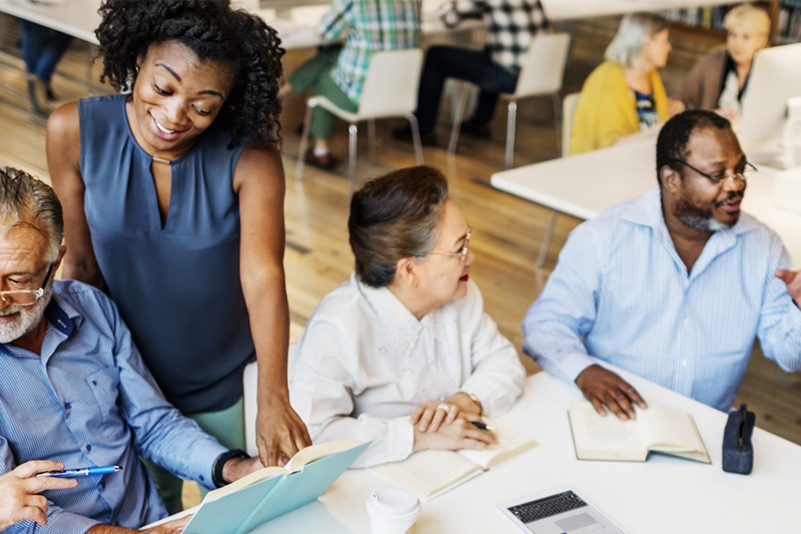 A group of diverse elderly individuals engaged in a discussion around a table with a laptop, notebooks, and documents in a library.
