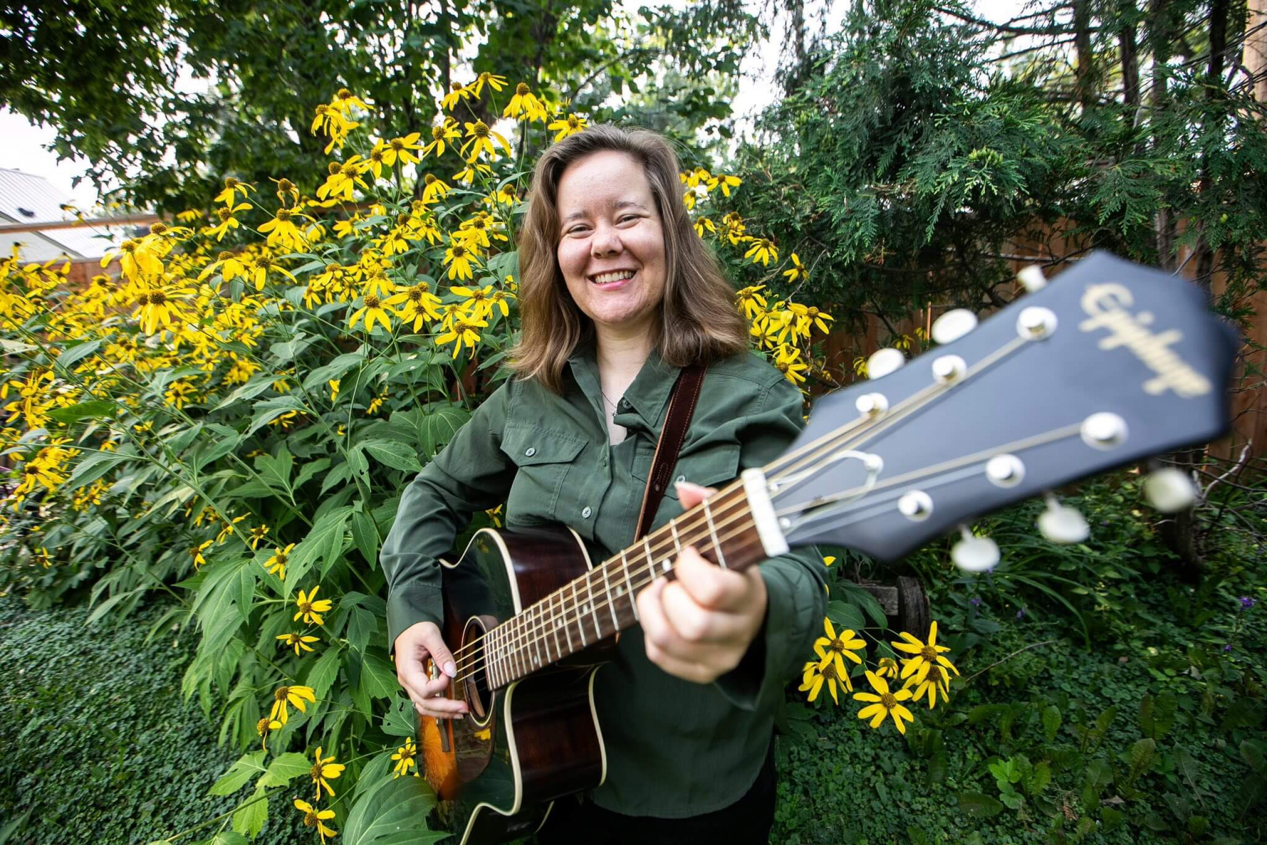 A smiling woman stands in front of yellow flowers holding a guitar.