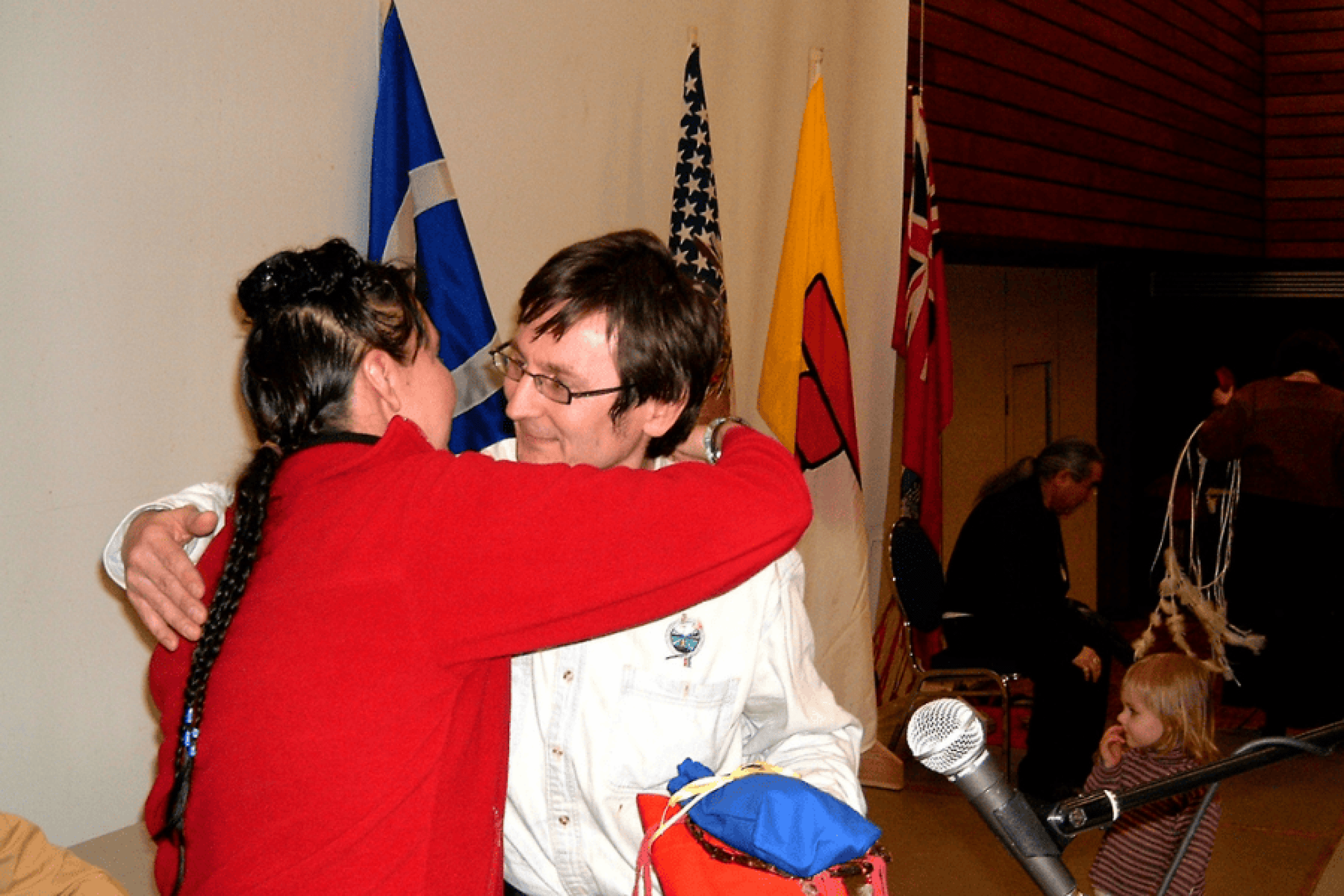 Two participants embrace during a moment of recognition at an Elders and Traditional Peoples Gathering, with flags displayed in the background.