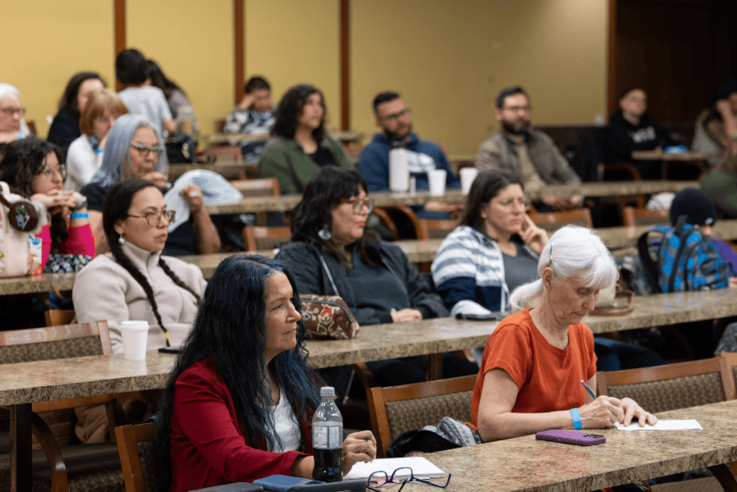 A group of people seated in a lecture-style room, listening and taking notes during a presentation.