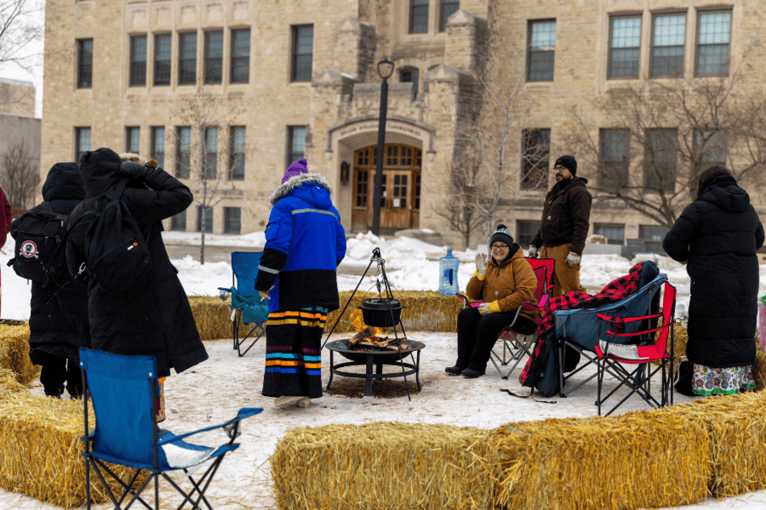 People gathered outdoors in winter around a small fire, seated on chairs and hay bales in front of a stone building.
