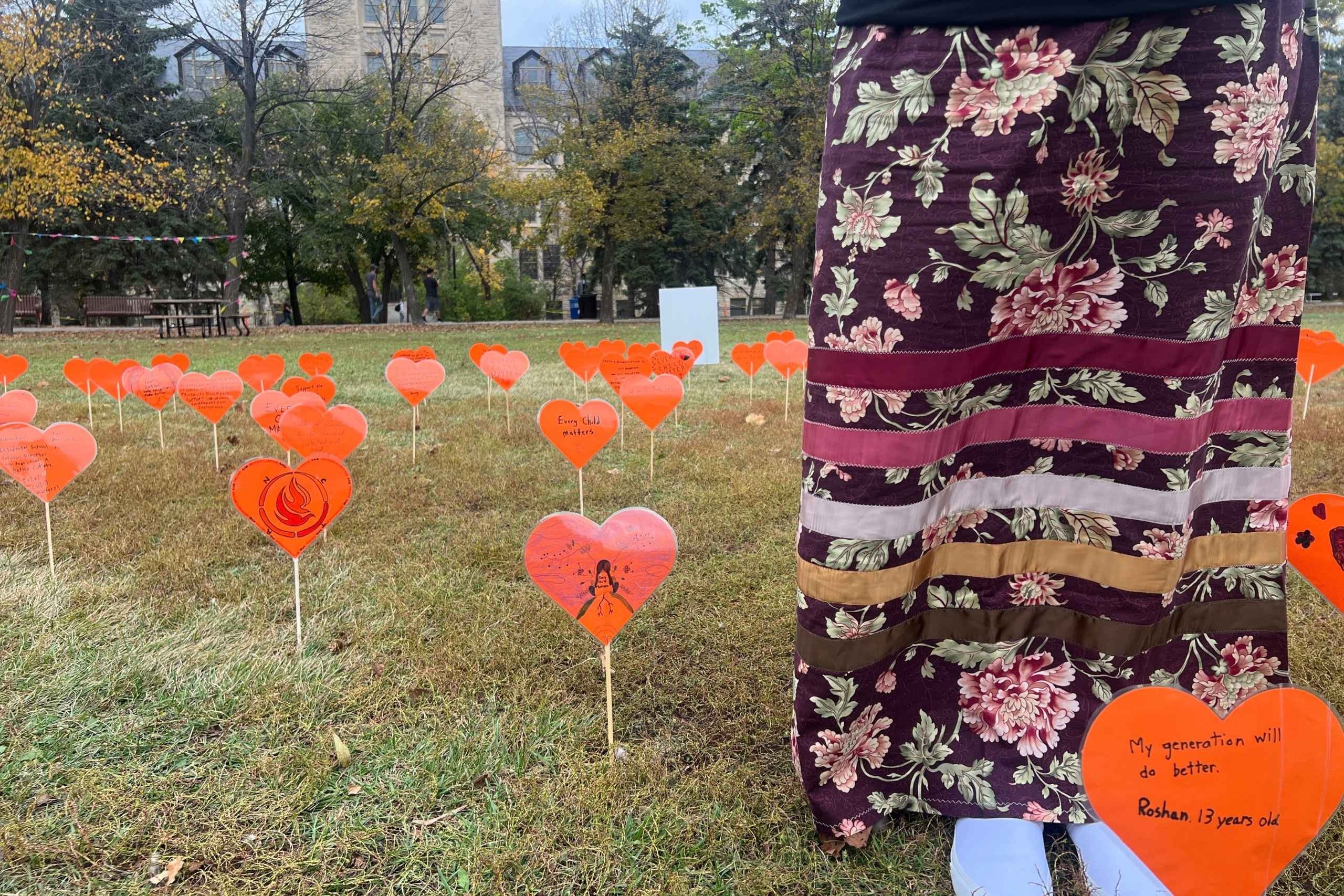 A person in a ribbon skirt, pictured from the waist down, stands in a field of orange hearts. The hearts feature messages of truth and reconciliation.