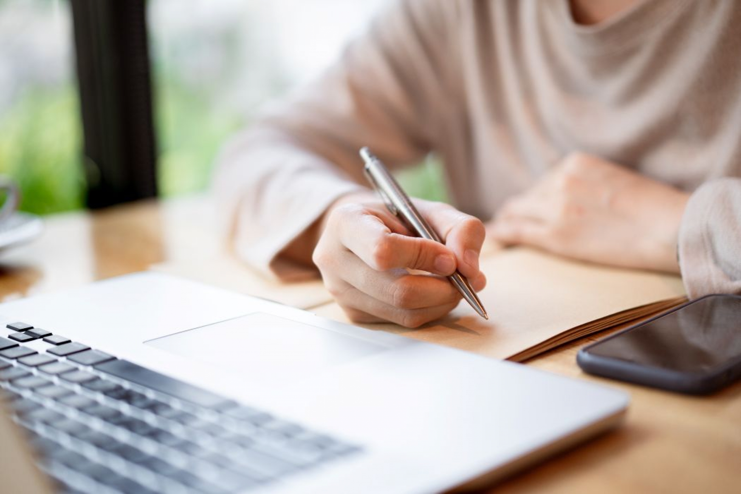 A person sits at a desk. They are about to write on a piece of paper with a pen. On the desk is a laptop and a smartphone.