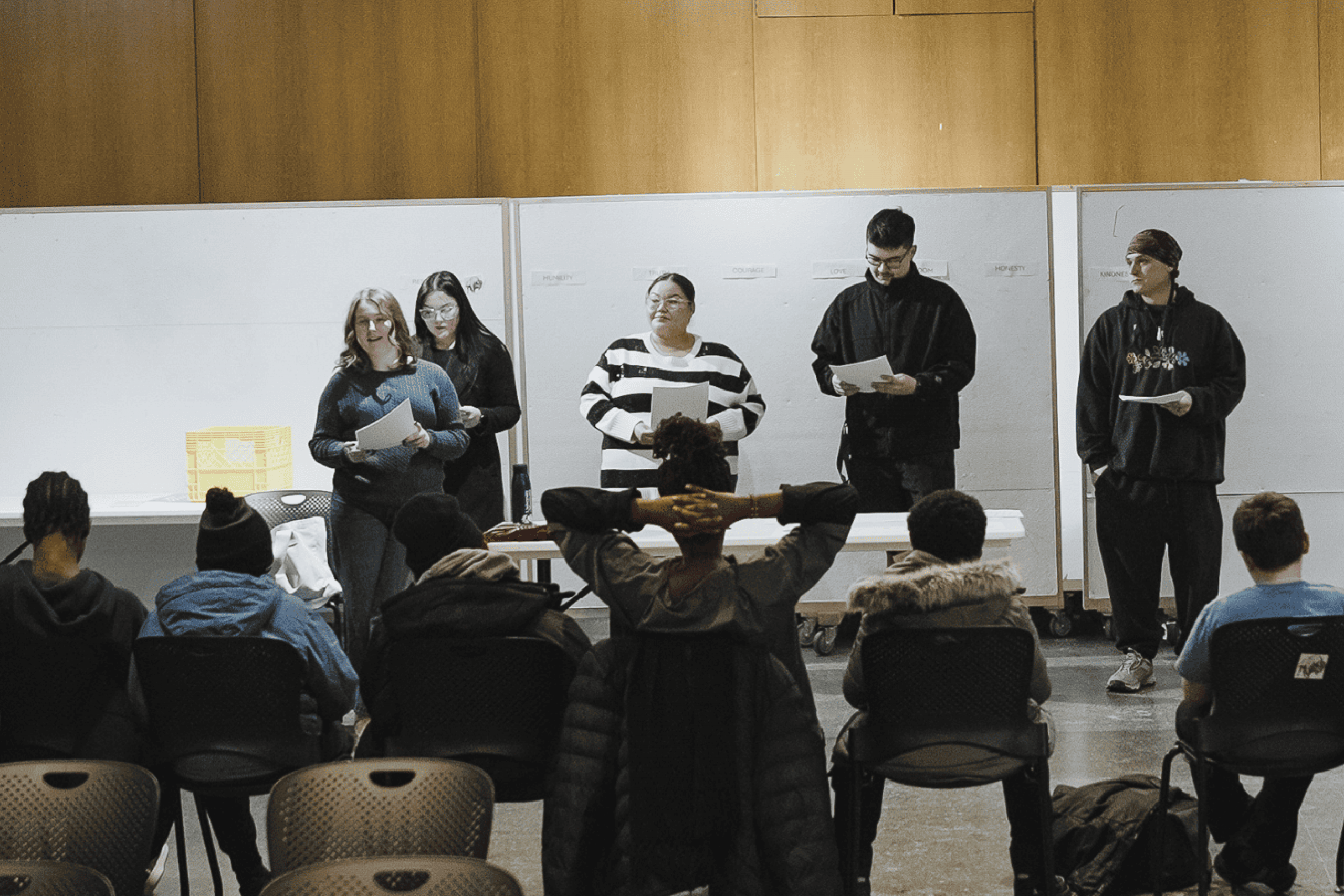 Students present to an audience in a classroom, reading from notes at the front of the room.