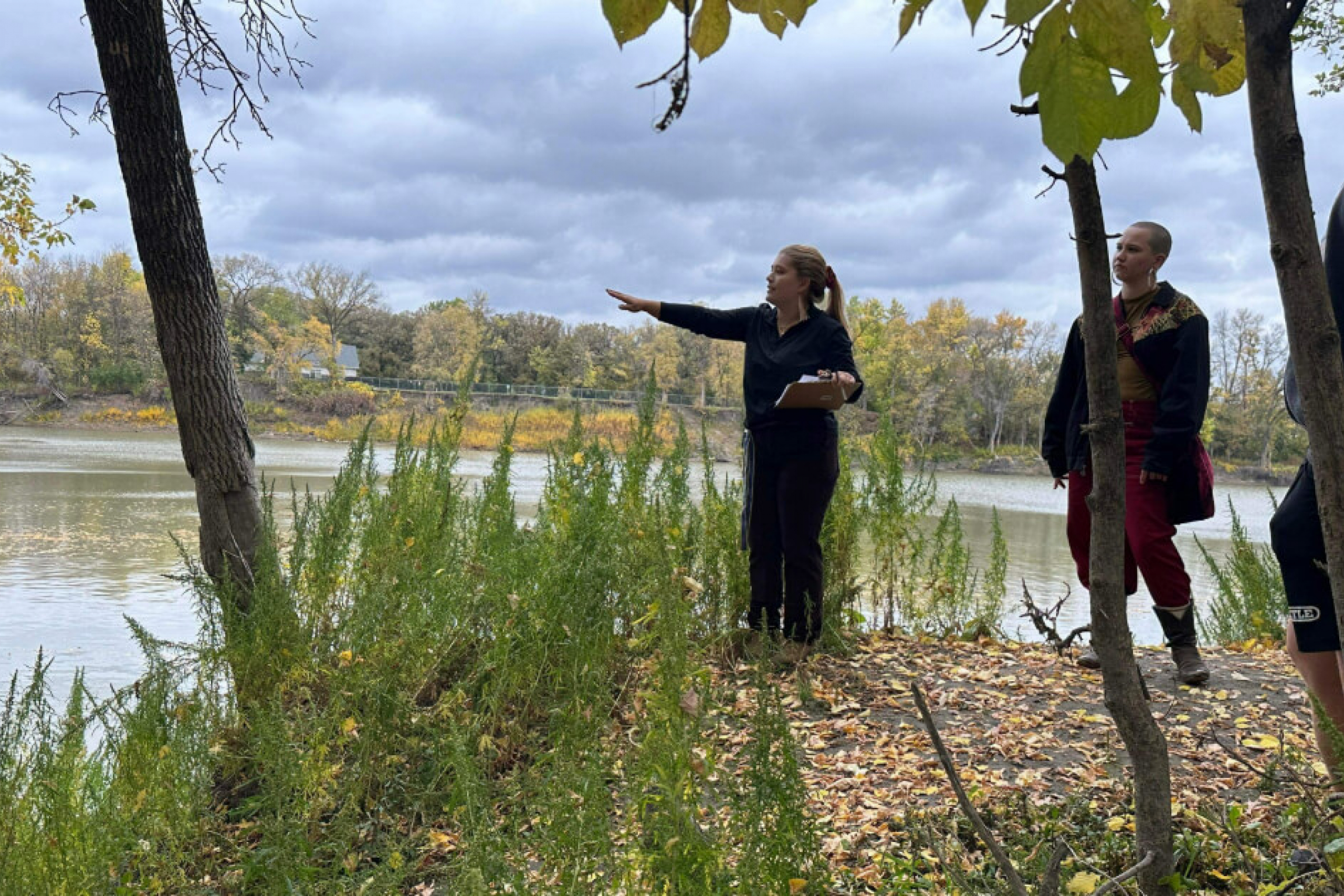 Three people standing on a riverbank with trees and water around them.