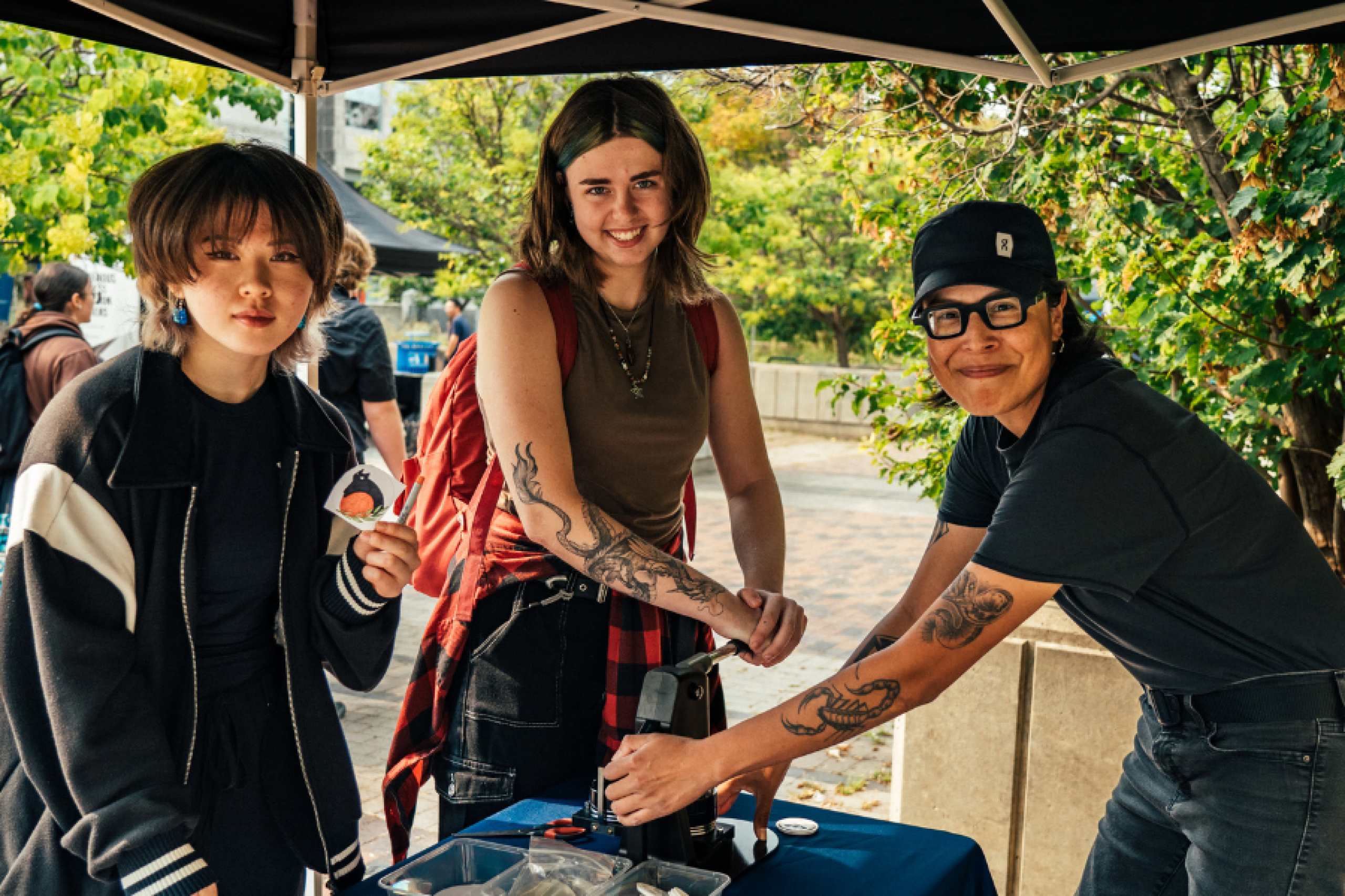 Three smiling students at a button-making booth during an outdoor event, enjoying the creative activity under a canopy.