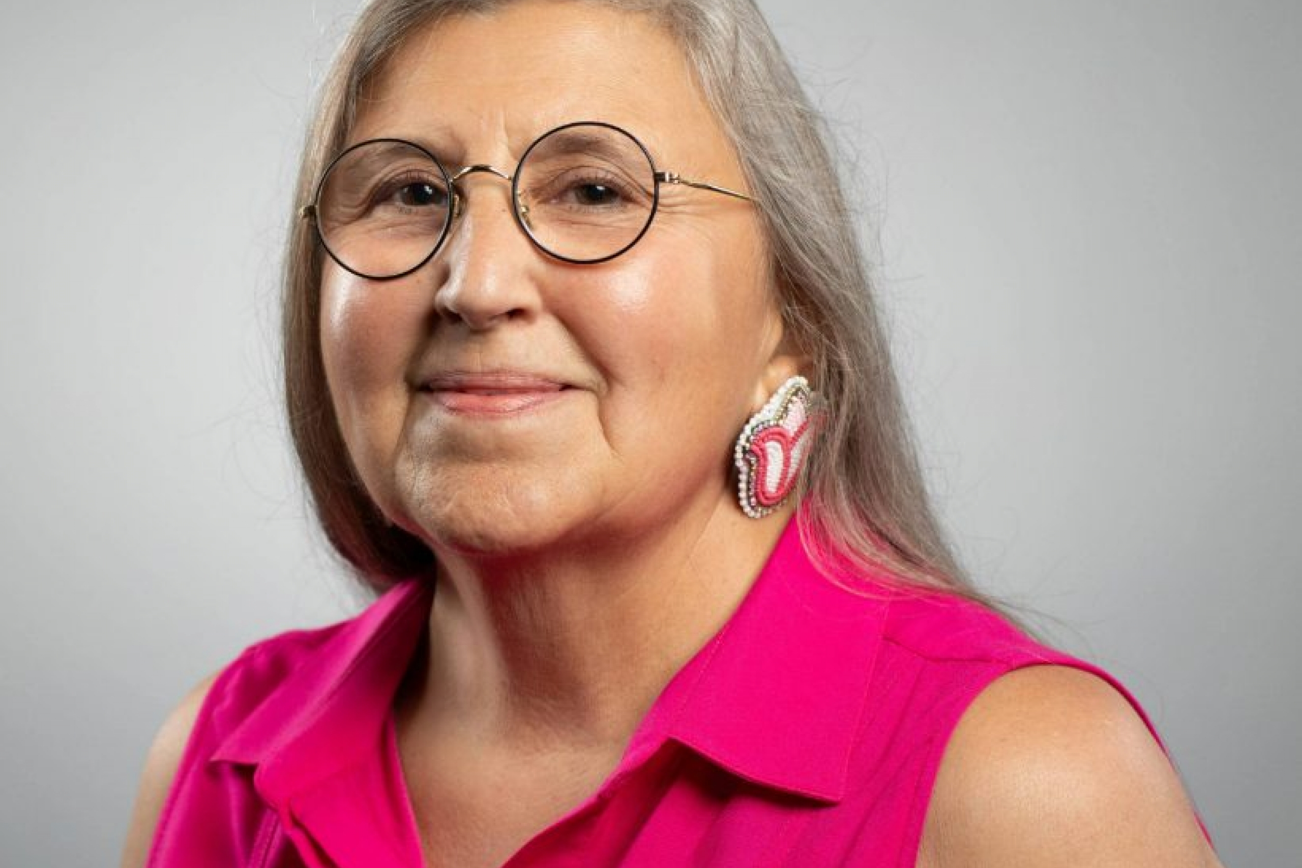 Headshot photo of Pahan PteSanWin. She is wearing glasses, facing the camera and smiling. She has long silver hair and is wearing a pink shirt, set against a grey background.