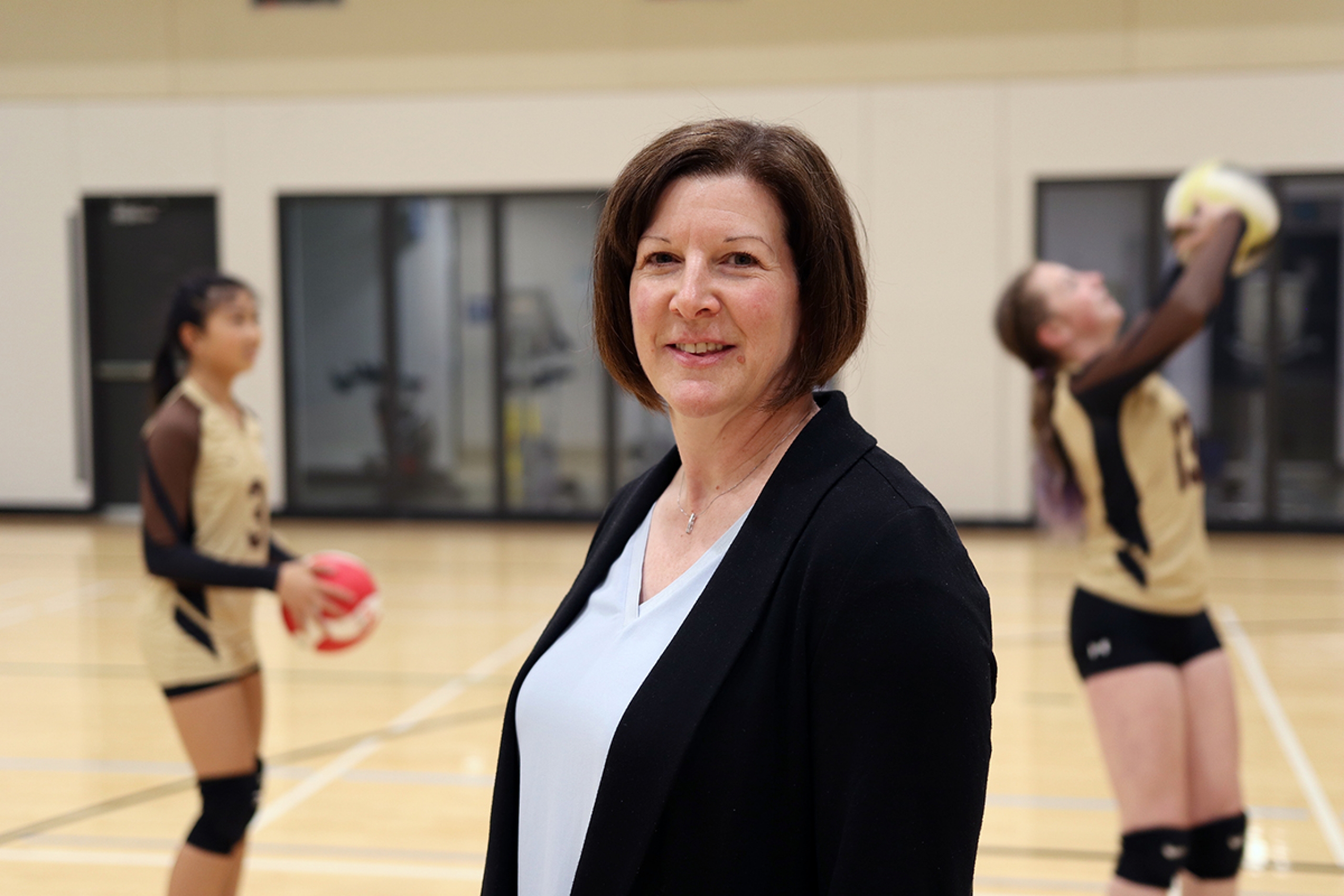 Joanne Parsons in a gymnasium with two teenage girls playing volleyball behind her.