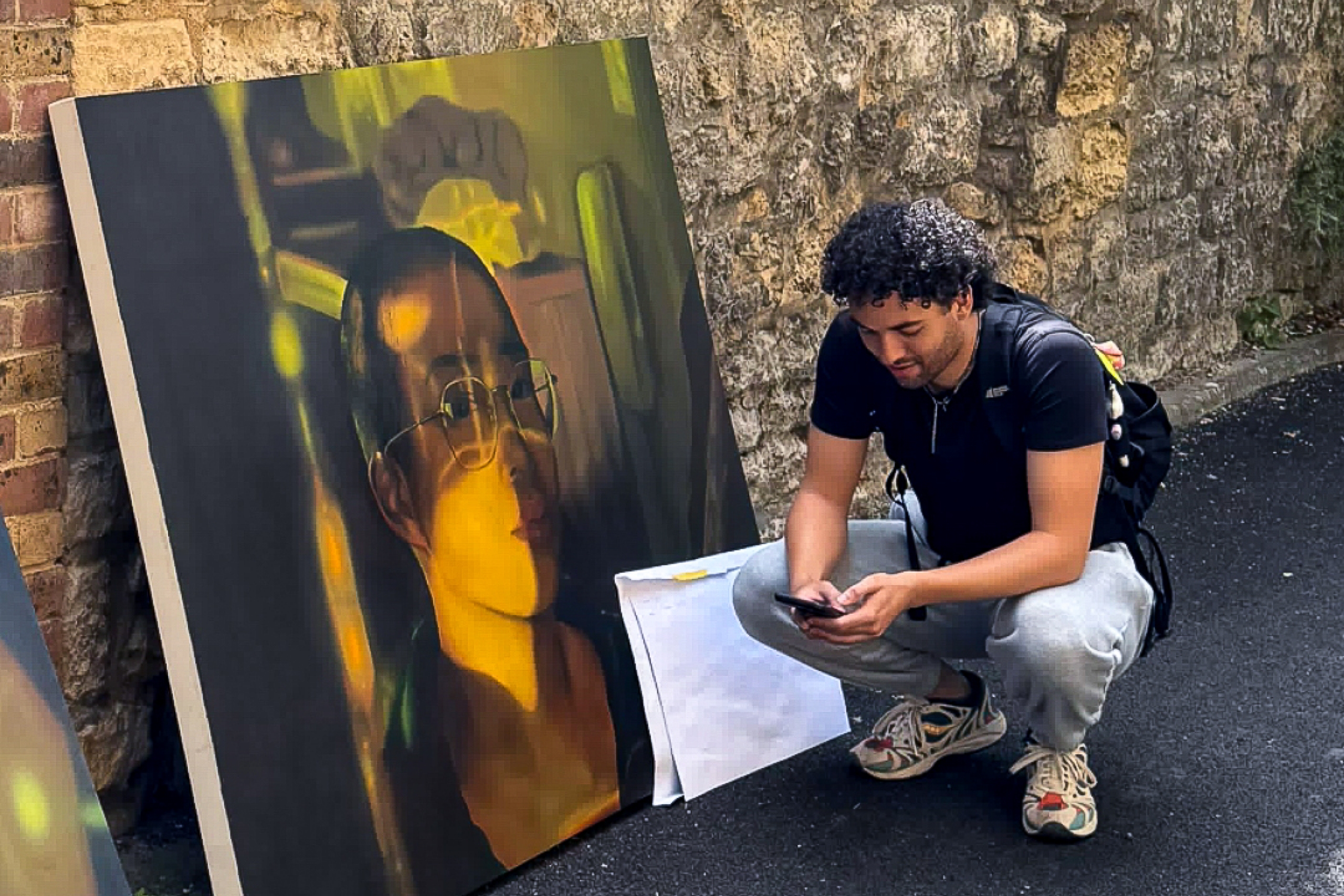Artist Joel Nichols crouches beside their oil painting In Our Borderlands against a stone wall.