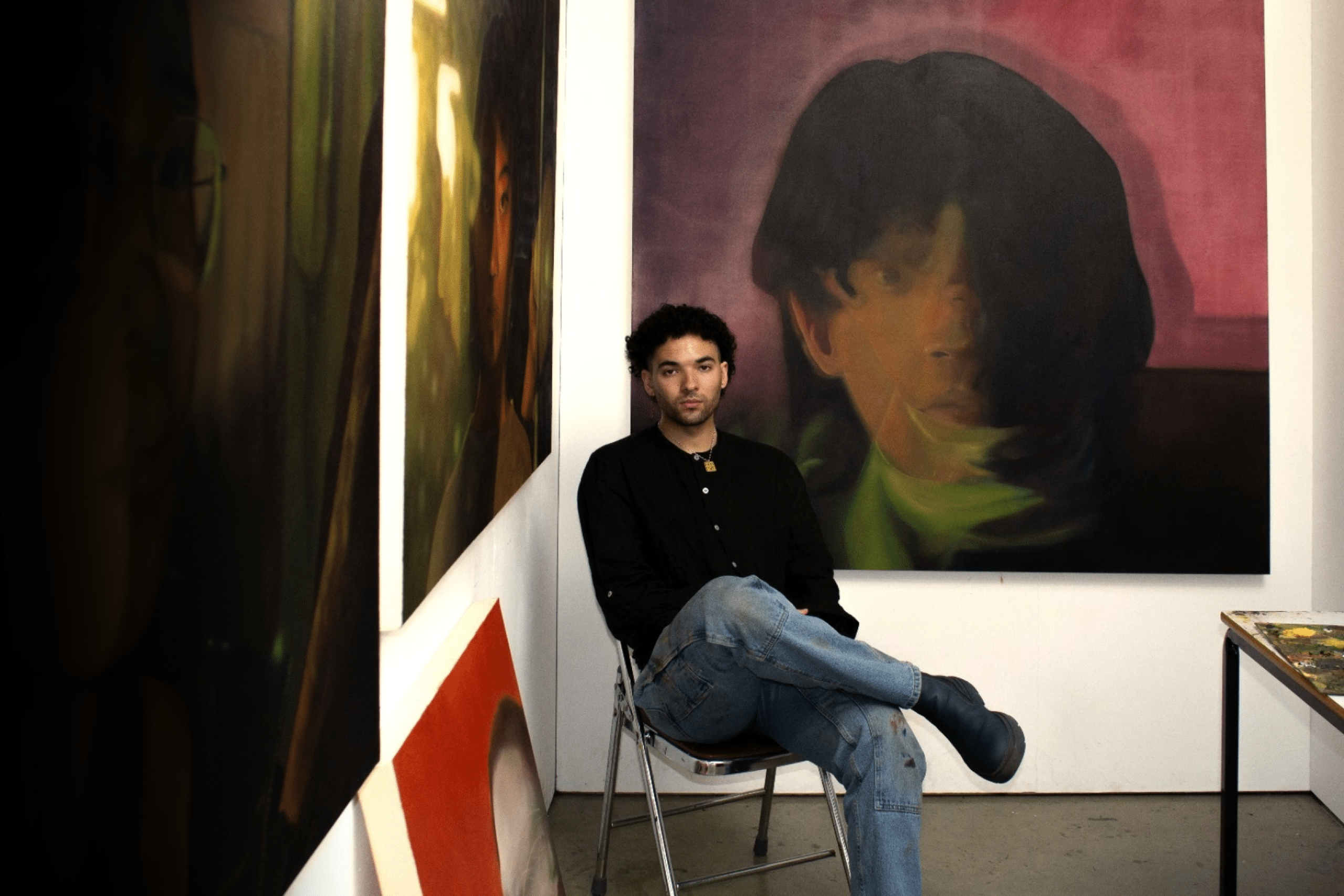 Joel Nichols sits in a studio surrounded by large-scale portrait paintings, with a paint-covered worktable nearby.