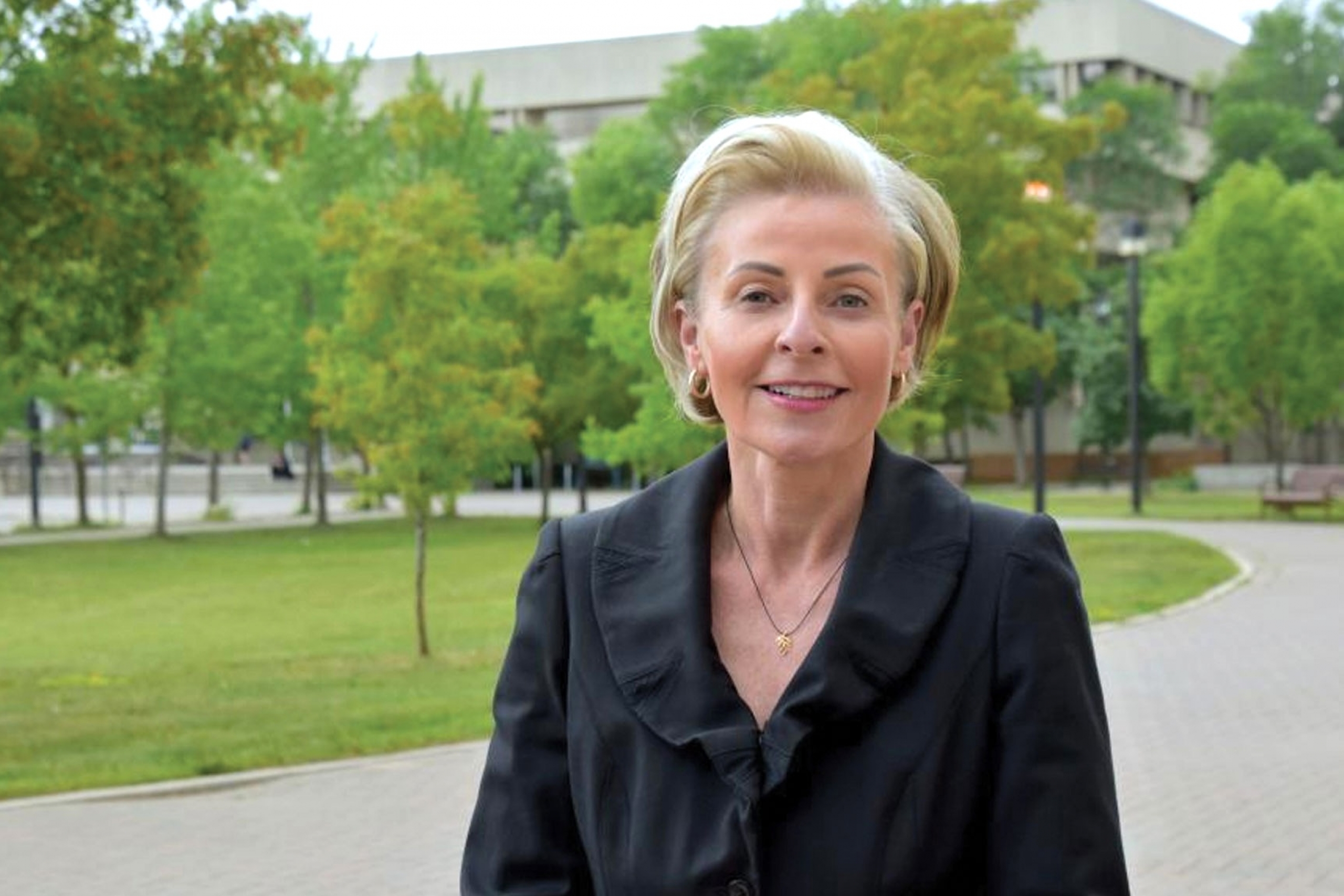 Krystyna Koczanski wearing a black blouse with ruffles with trees background smiling at the camera.