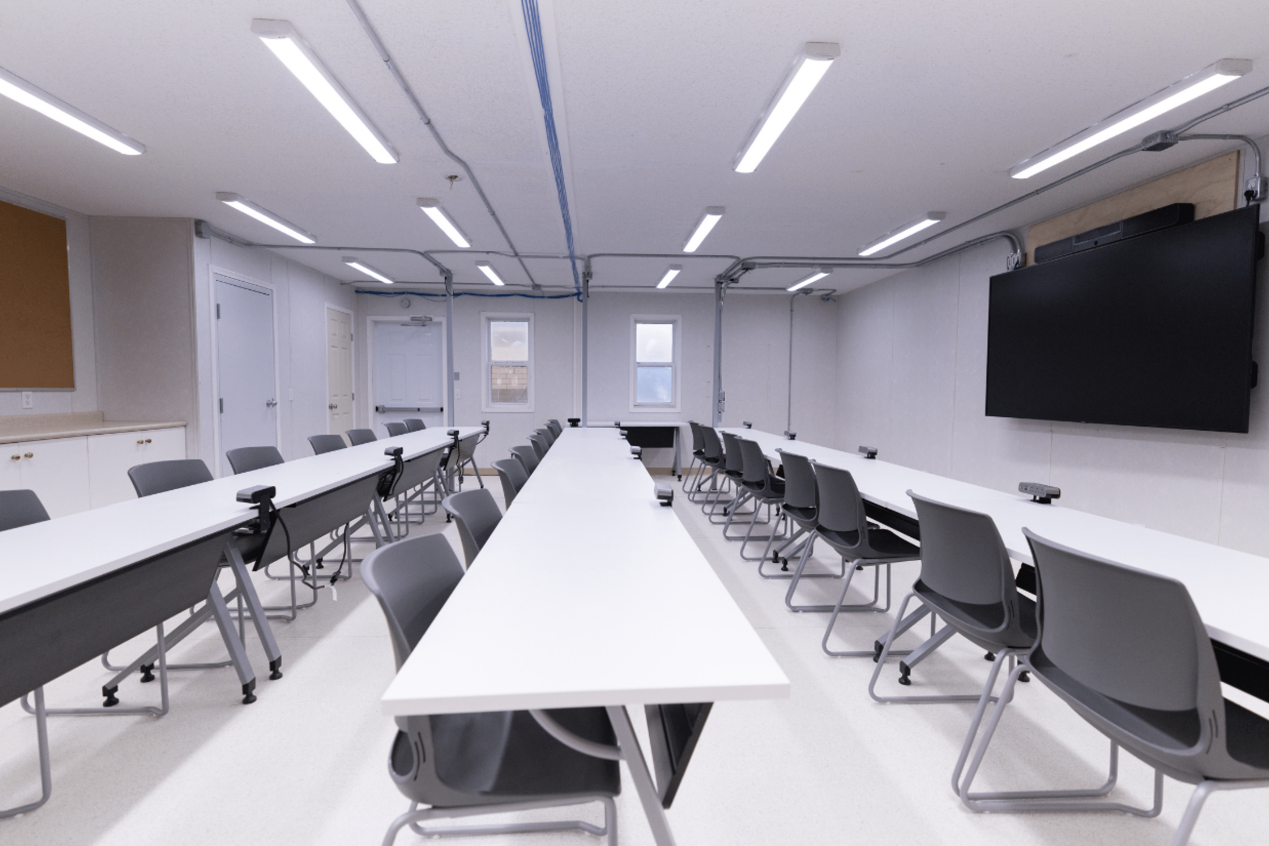 Modern classroom with long tables, gray chairs, and a wall-mounted screen.