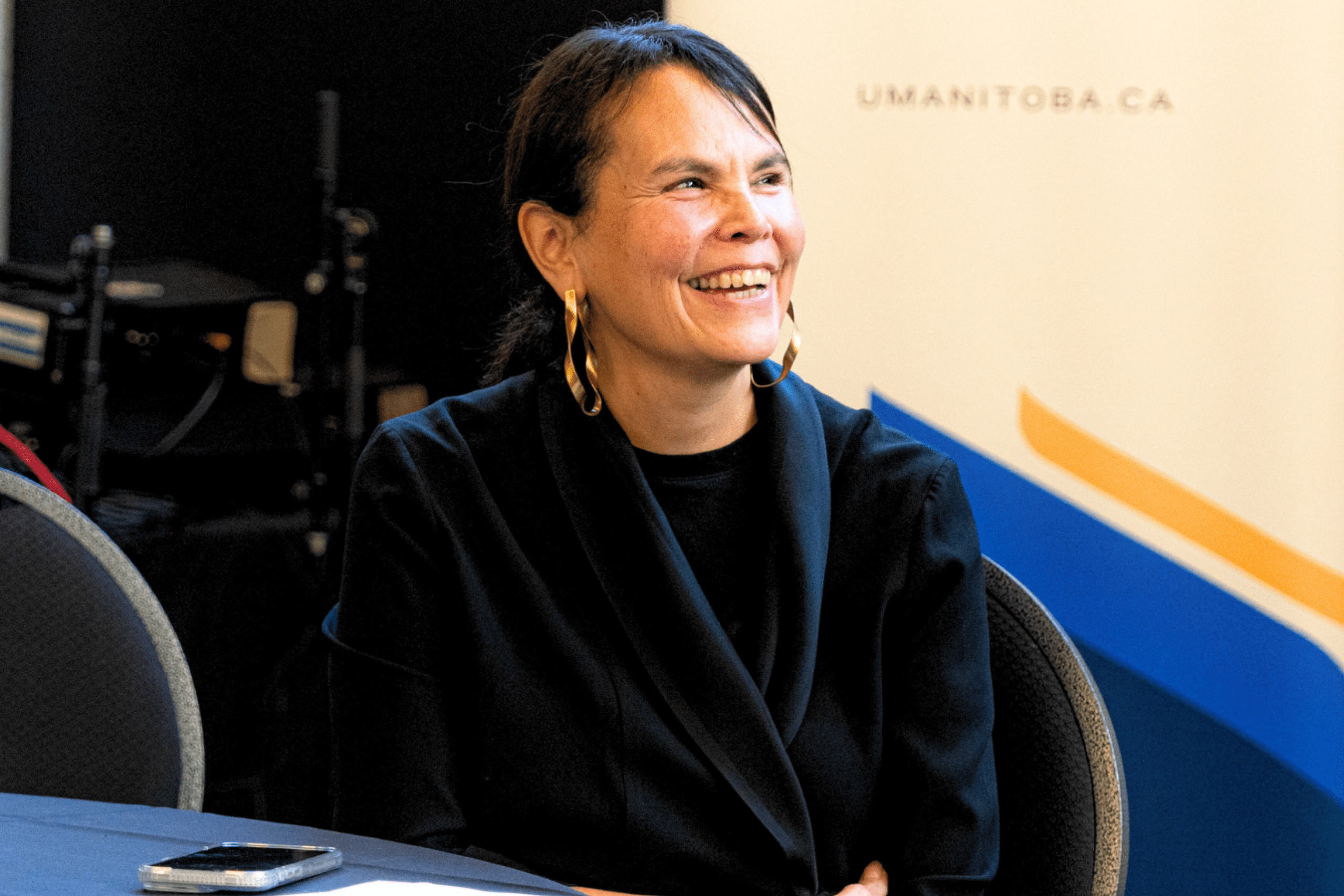 A woman seated at a table at a university event, smiling warmly, with a mobile phone placed in front of her.