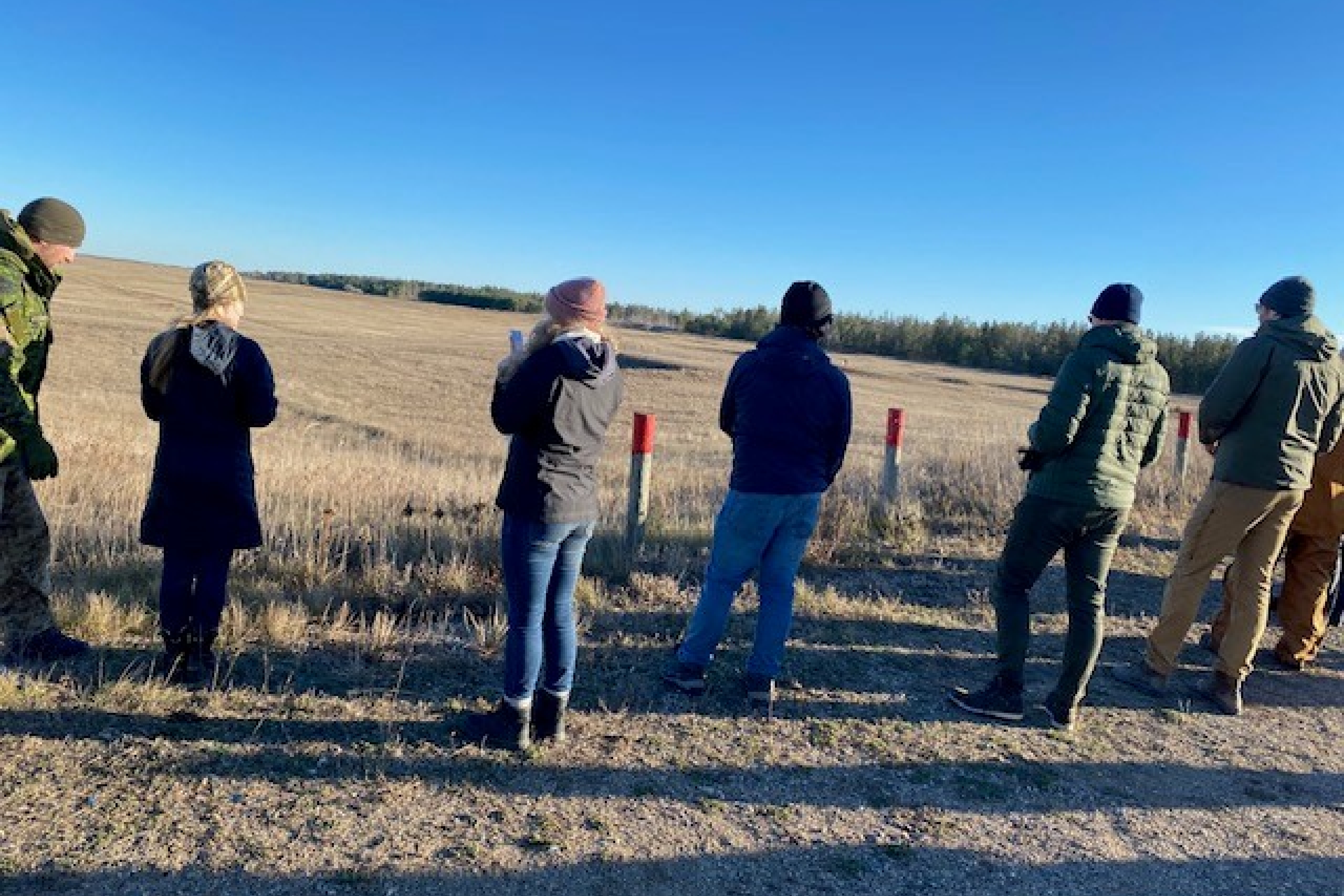 Students, with their backs to us, standing in a field, receiving instruction from military personnel in full fatigues.