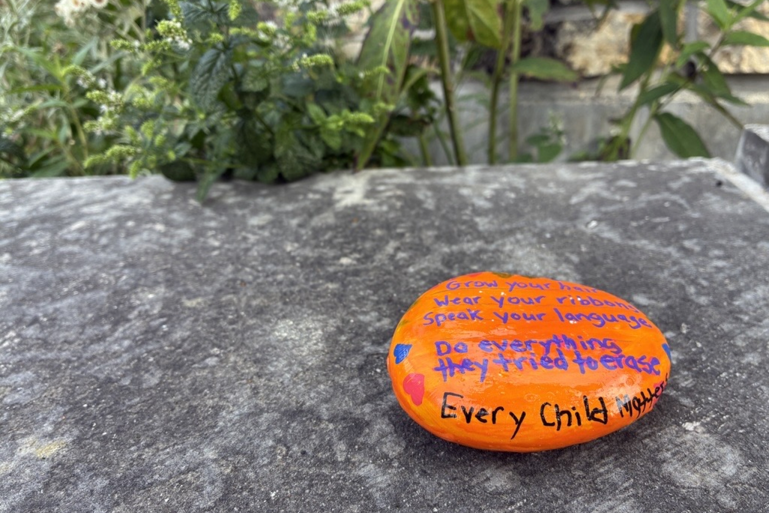 An orange-painted rock is placed outside on a stone ledge with green plants in the background.