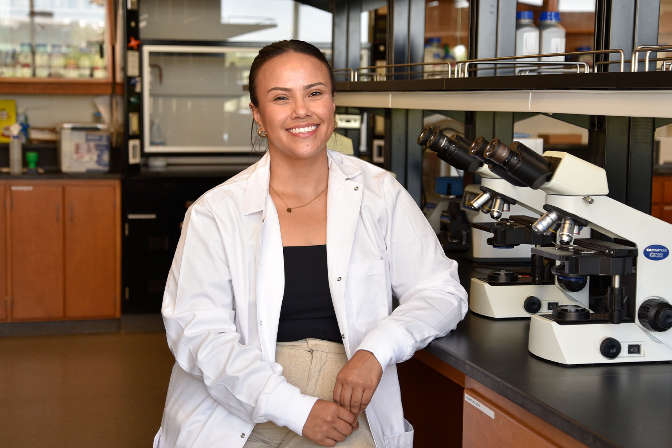 Rebecca Asham wearing a lab coat and smiling at the camera beside a microscope.