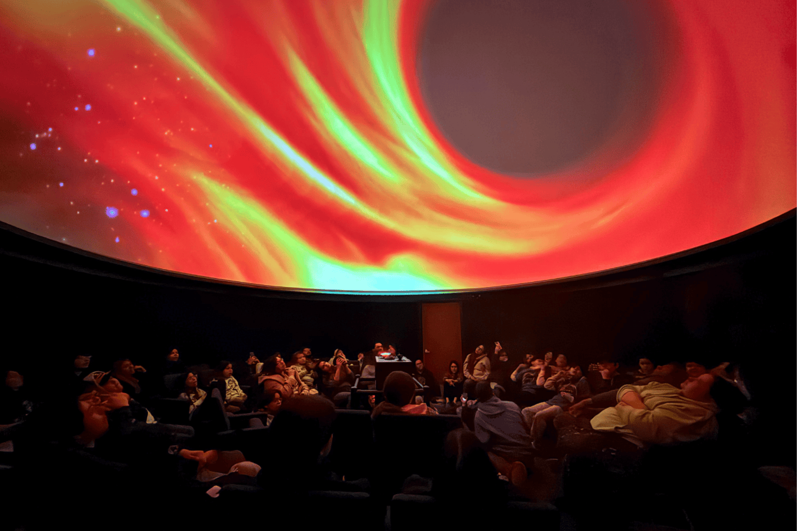 A group of attendees seated in a dome theatre, watching a vivid projection of a swirling cosmic scene resembling a black hole.