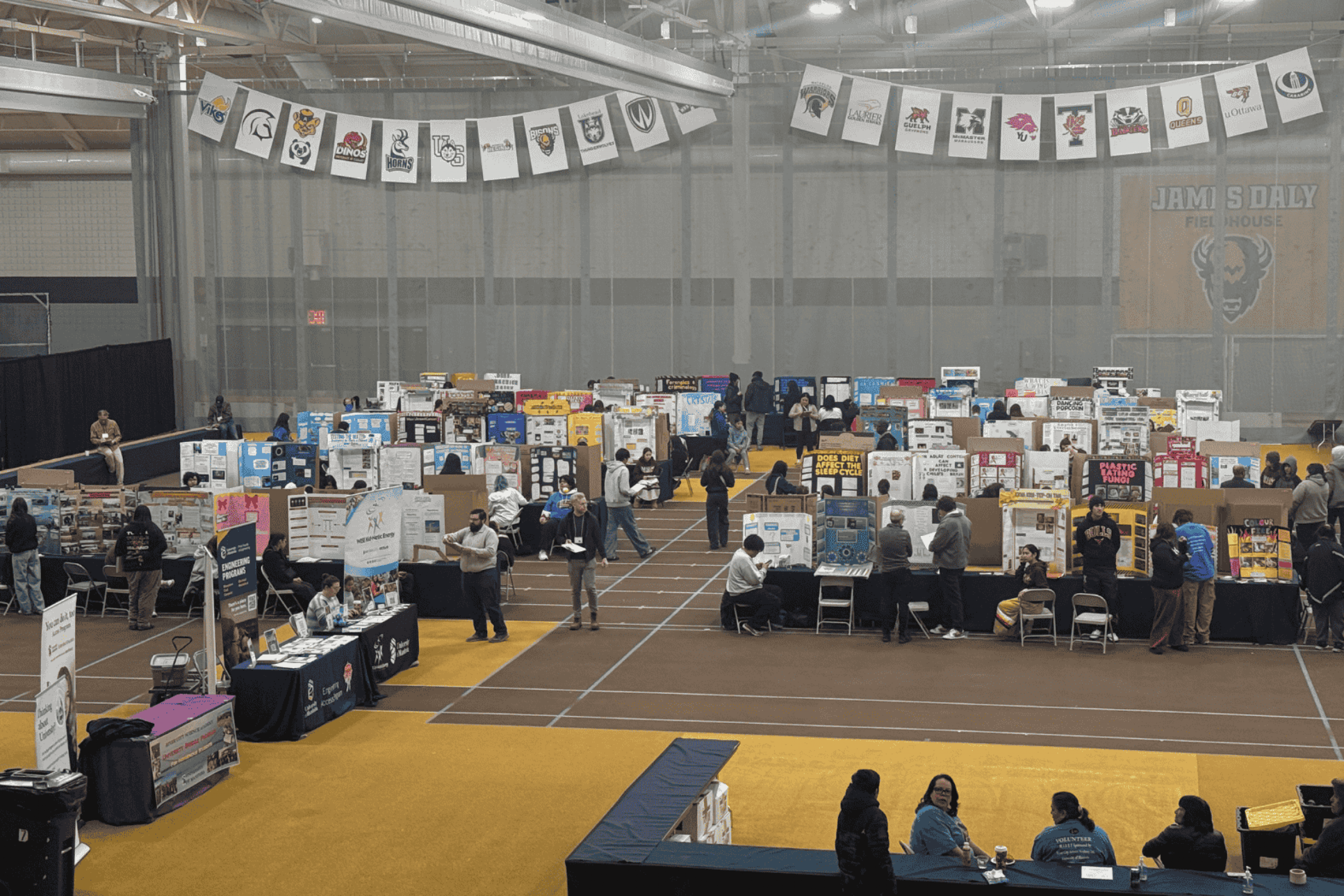 Wide view of a science fair with student project displays and attendees interacting in a large indoor space.