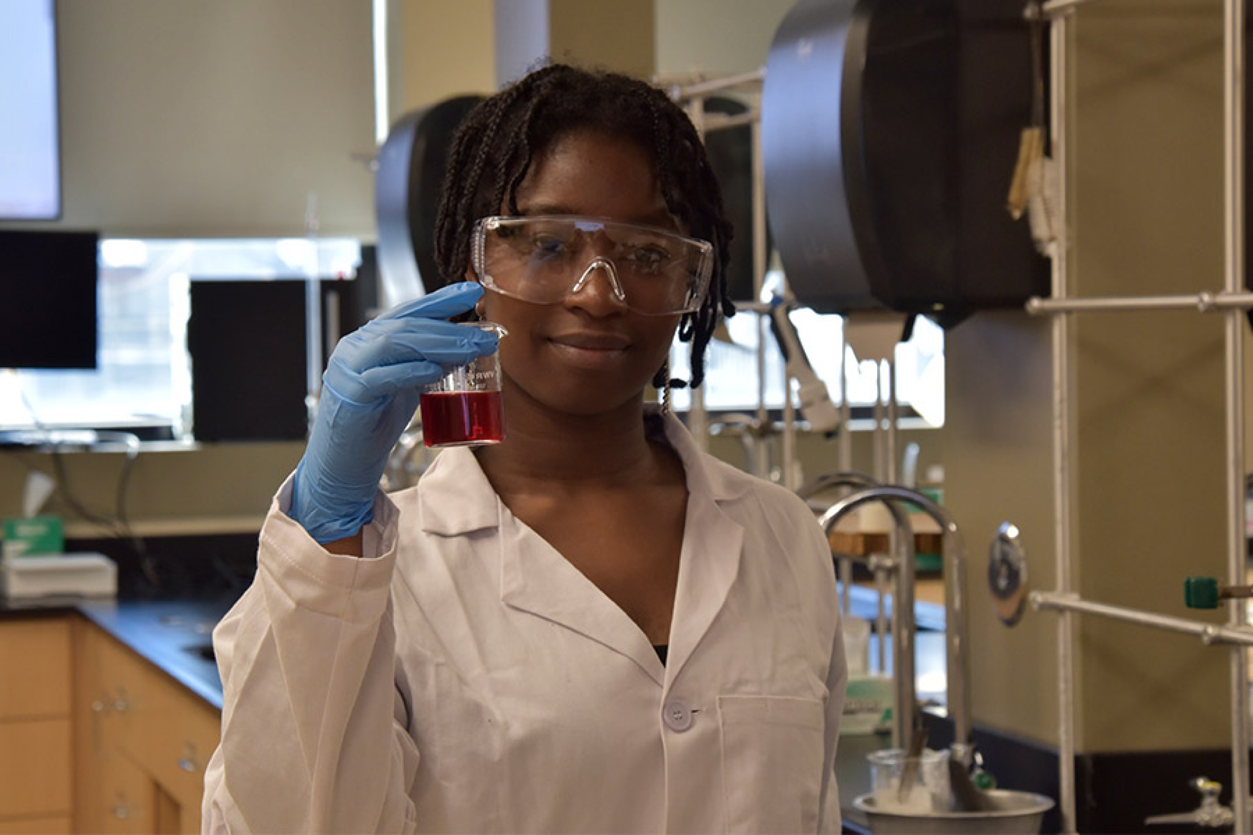 Seun wearing a safety goggles and a labcoat holding a test tube smiling at the camera.