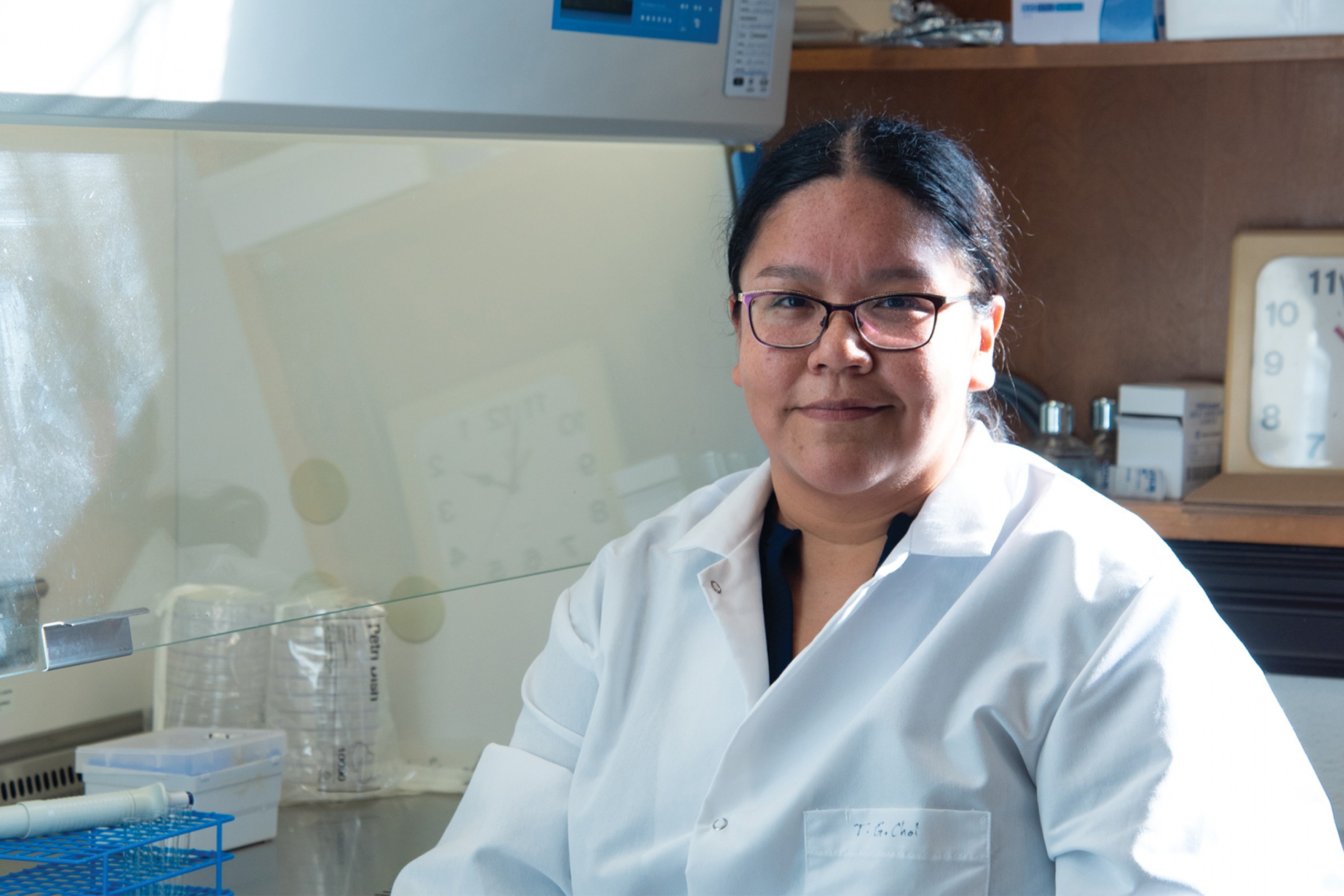 Shana wearing a lab coat beside the biosafety cabinet smiling at the camera.