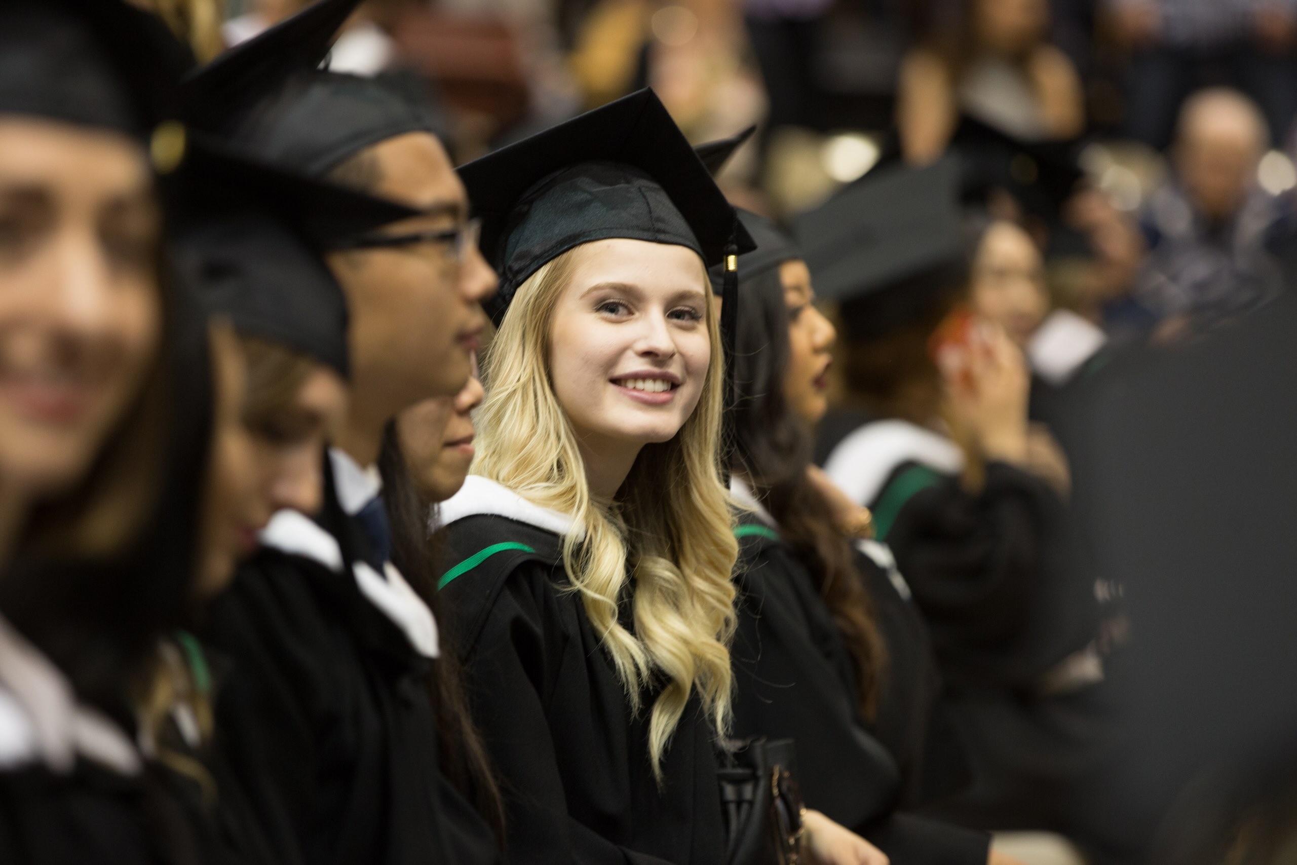 Young smiling woman in black grad cap and gown sits with other graduands. 