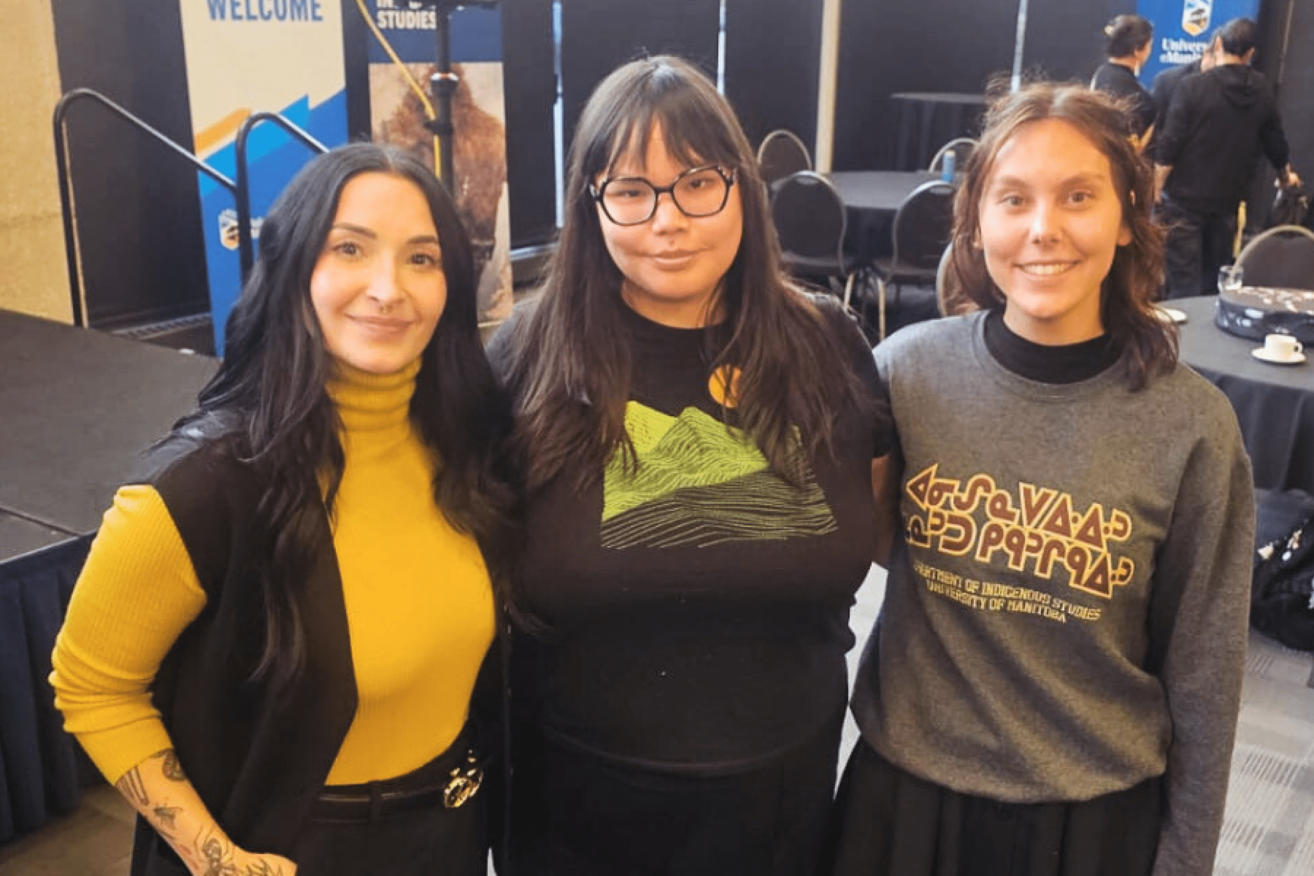Three women standing together and smiling at an indoor Indigenous community event.