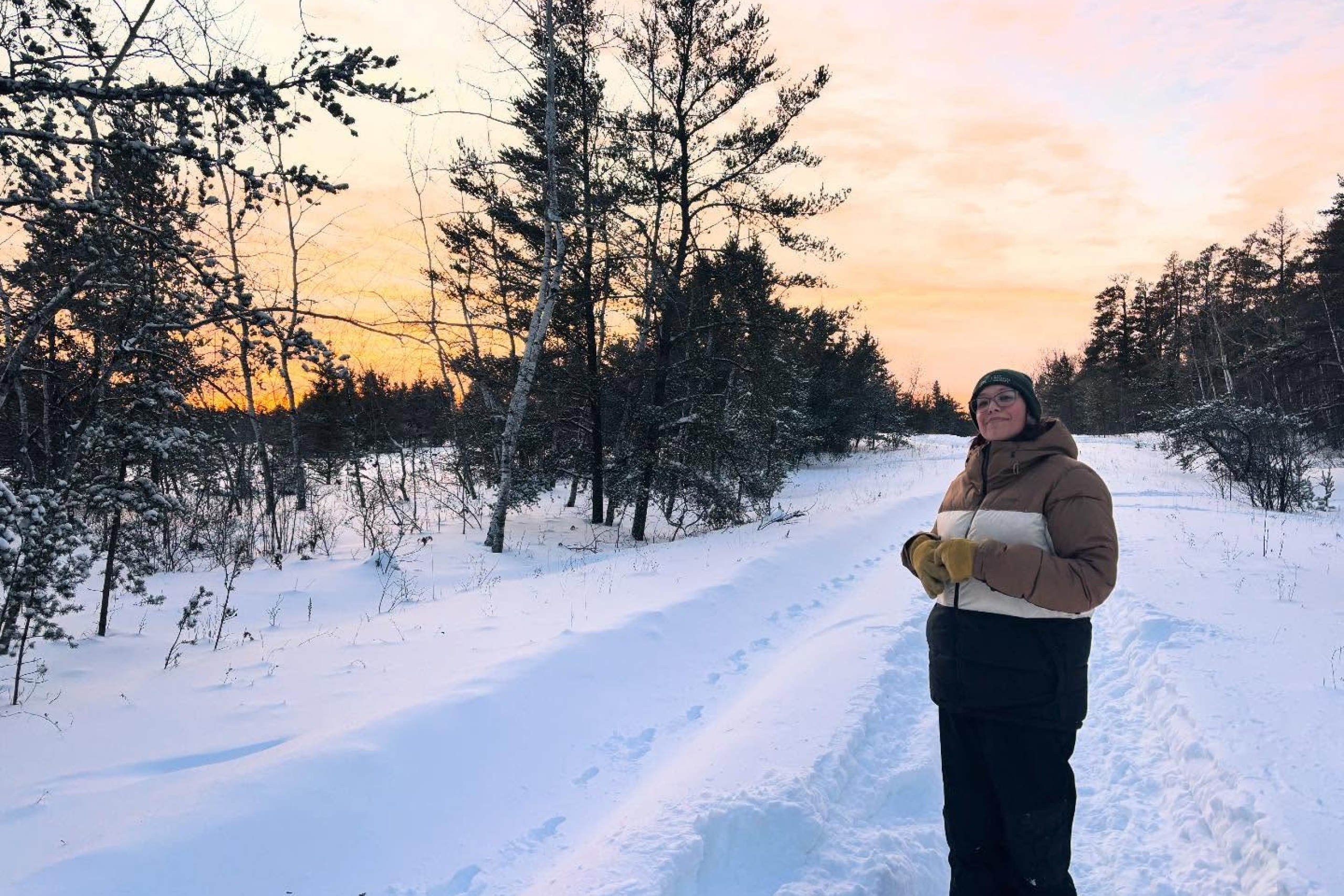 Taylor smiles with a sunset in the background, snow on the ground and trees on the horizon.