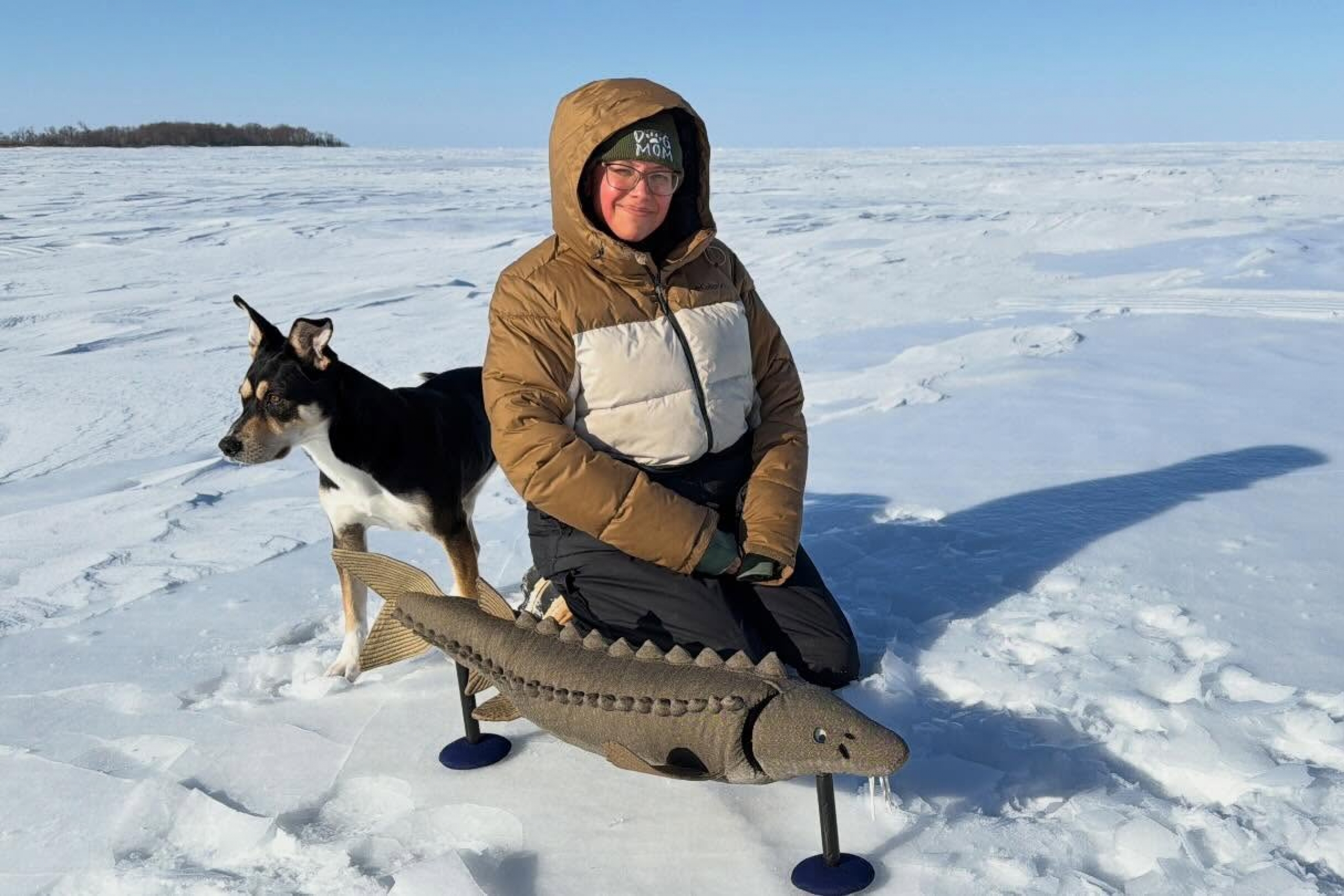 female kneels on snow over frozen lake with her dog beside her and sturgeon fish puppet in front.